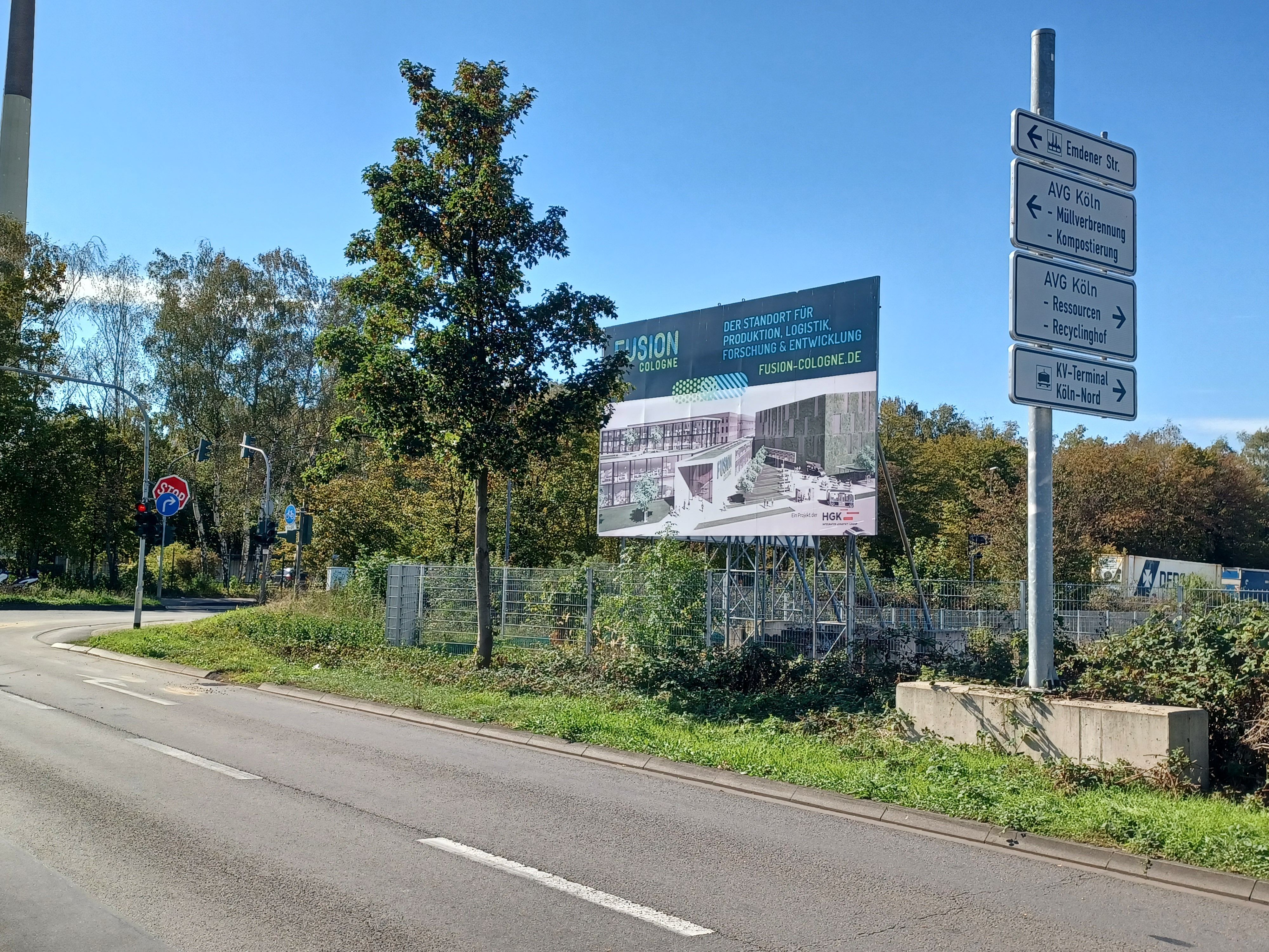 Das Foto zeigt eine Straße mit Verkehrsschildern und einer Werbetafel vor einem umzäunten, von Bäumen und Sträuchern umgebenen Gelände.