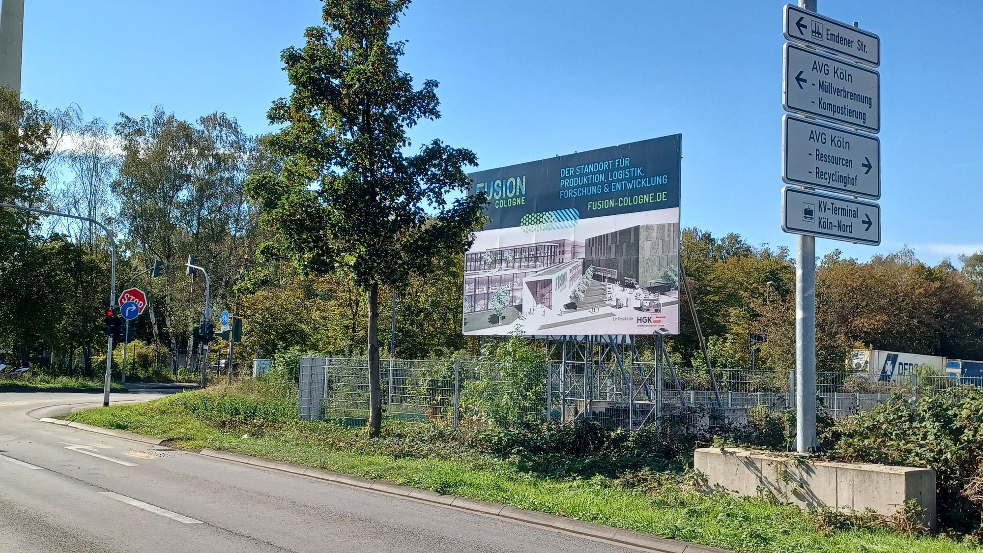 Das Foto zeigt eine Straße mit Verkehrsschildern und einer Werbetafel vor einem umzäunten, von Bäumen und Sträuchern umgebenen Gelände.