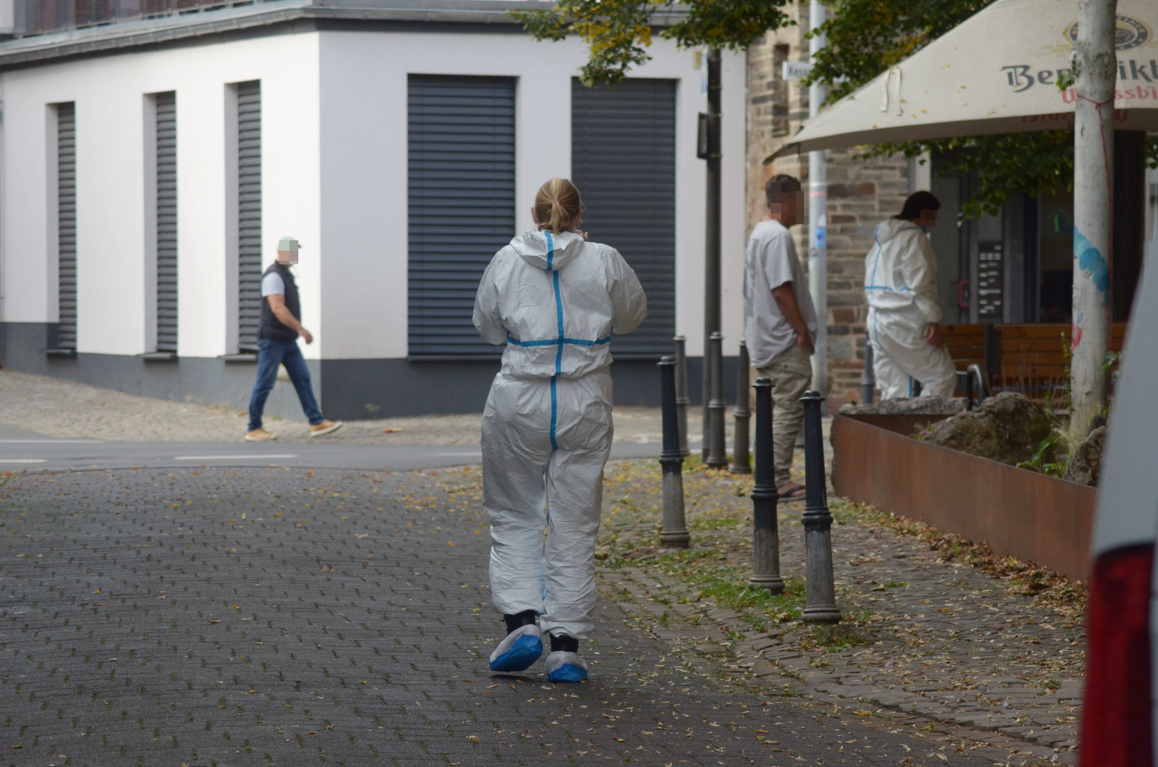 Das Bild zeigt zwei Einsatzkräfte in weißen Schutzanzügen sowie zwei Passanten.