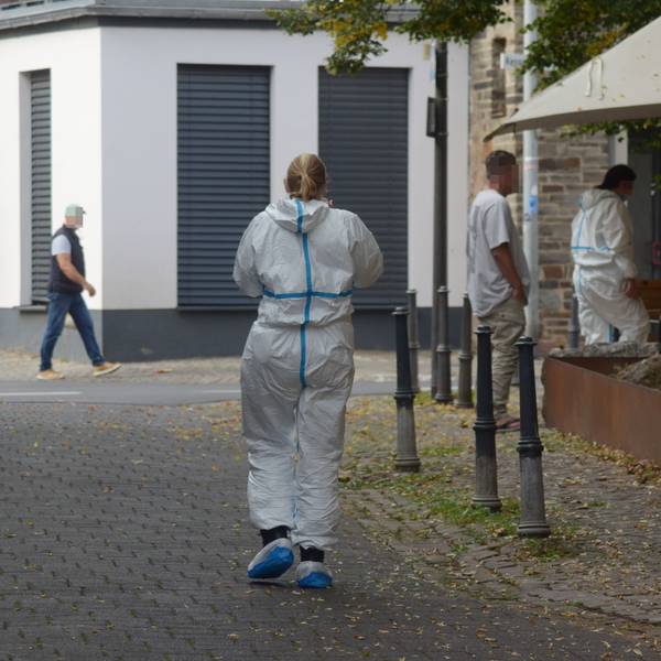 Das Bild zeigt zwei Einsatzkräfte in weißen Schutzanzügen sowie zwei Passanten.