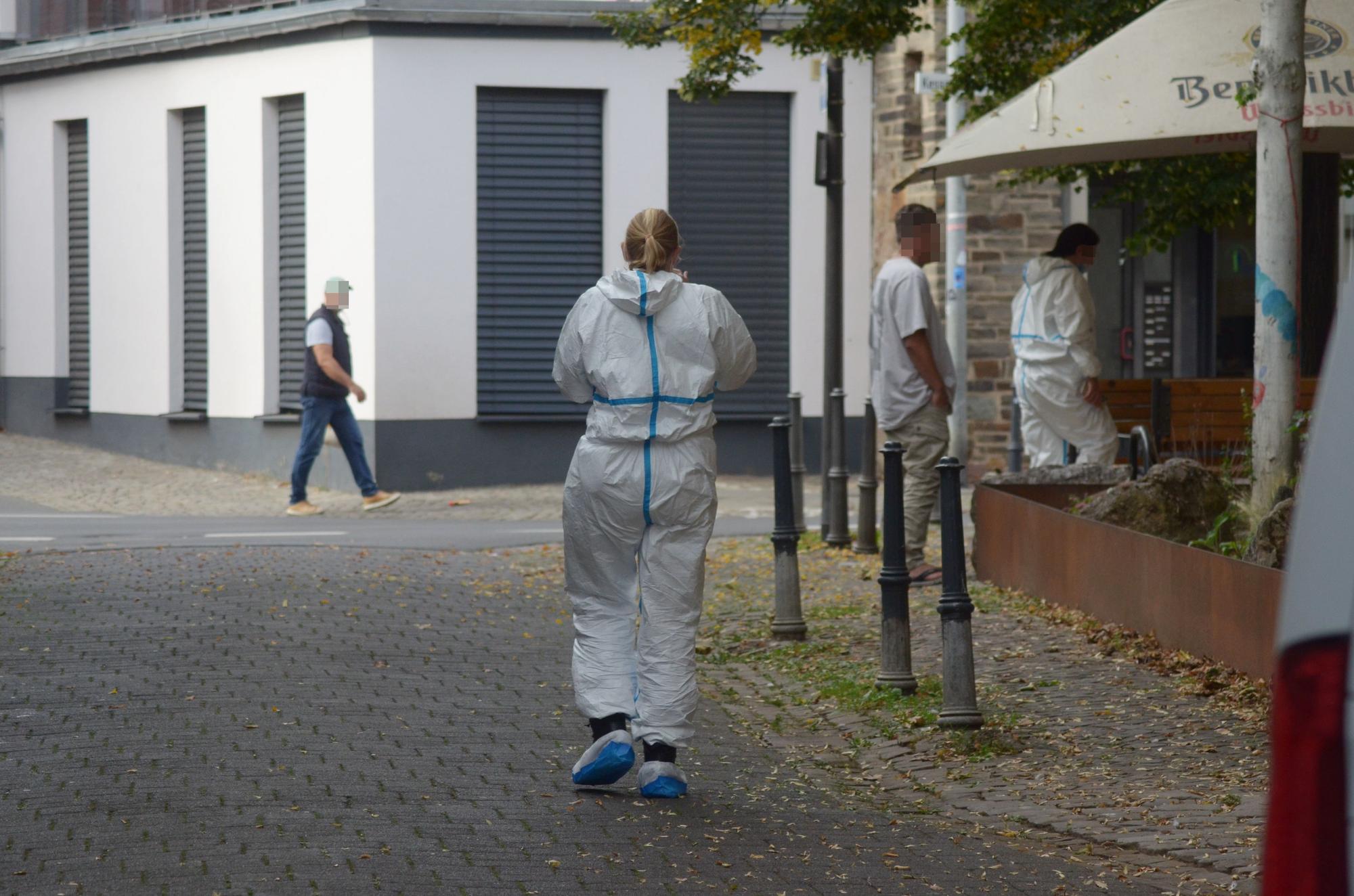 Das Bild zeigt zwei Einsatzkräfte in weißen Schutzanzügen sowie zwei Passanten.