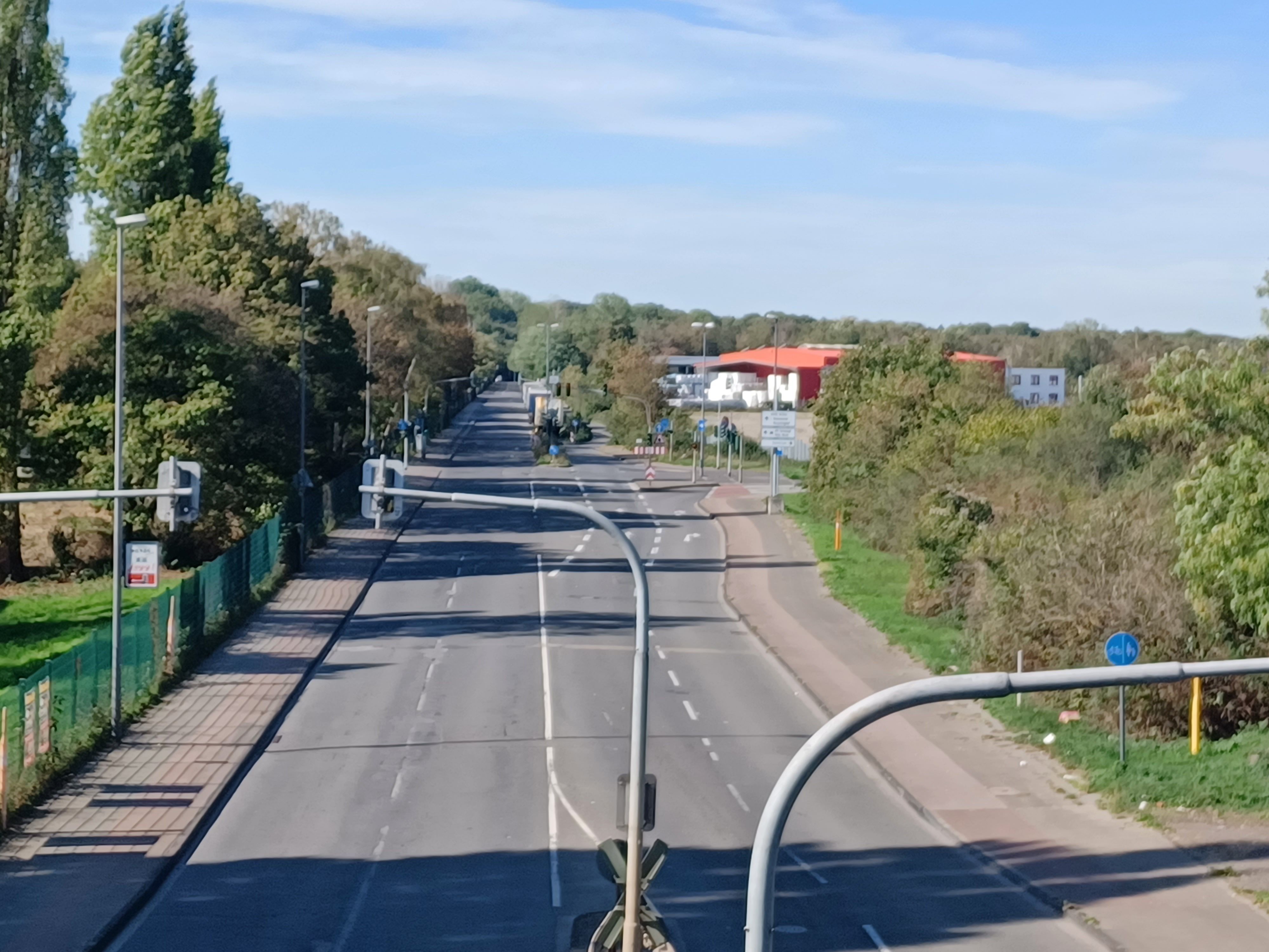 Das Foto zeigt eine mehrspurige Straße mit Gehwegen, Ampeln und Verkehrsschildern, die von Bäumen und Gebäuden gesäumt ist.