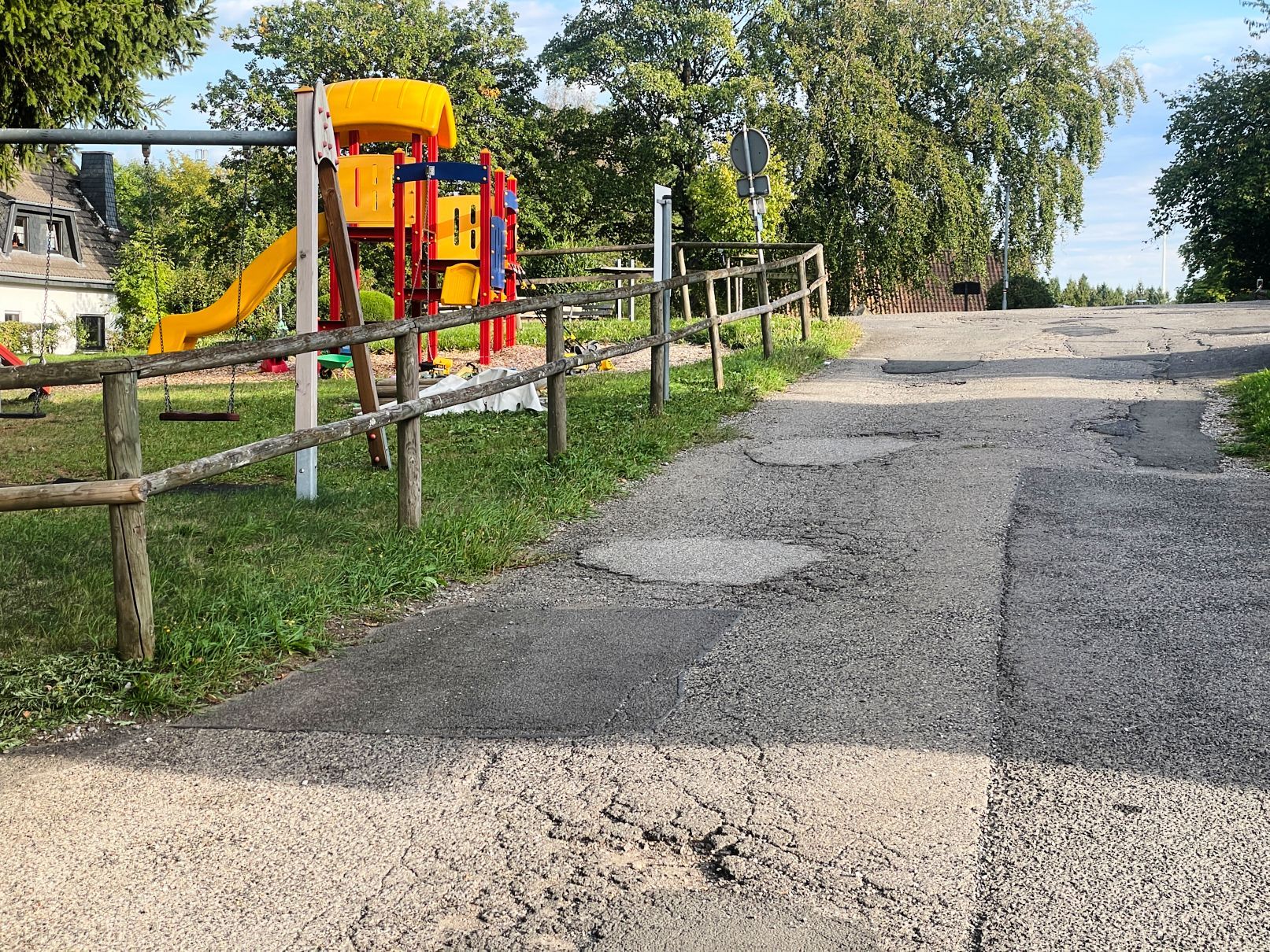 An einem Spielplatz führt eine mit etlichen Flickstellen versehene Asphalstraße vorbei.