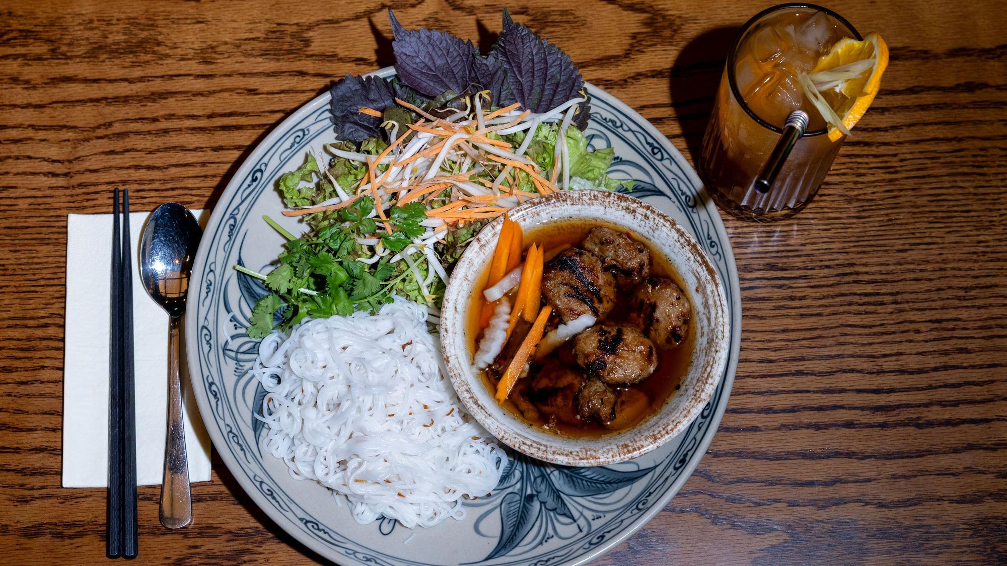 Bun Cha Hanoi: gegrilltes Schweinefleisch mit würzigen Hackfleischbällchen, Reisnudeln, Salat und Kräutern, angerichtet auf einem Teller, danben steht eine Limonade