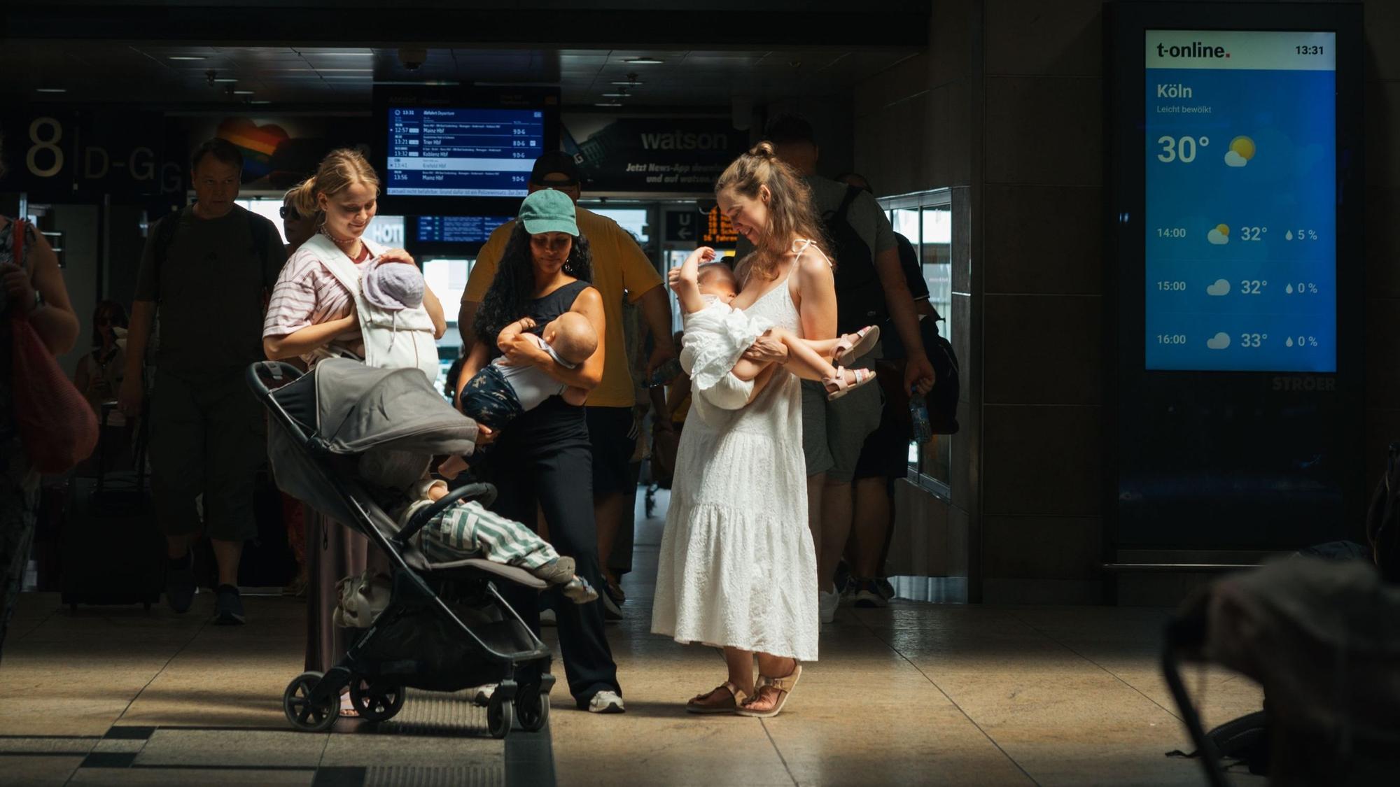 Drei Frauen stehen im Kölner Hauptbahnhof und stillen ihre Kinder.