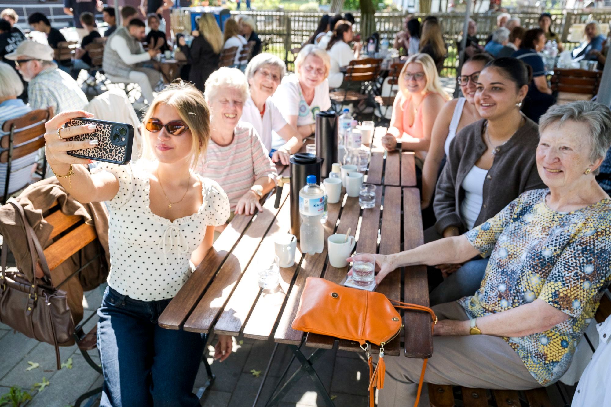 Junge und alte Frauen sitzen um einen Tisch im Gartenrestaurant; eine junge Frau nimmt ein Selfie mit der Gruppe auf.