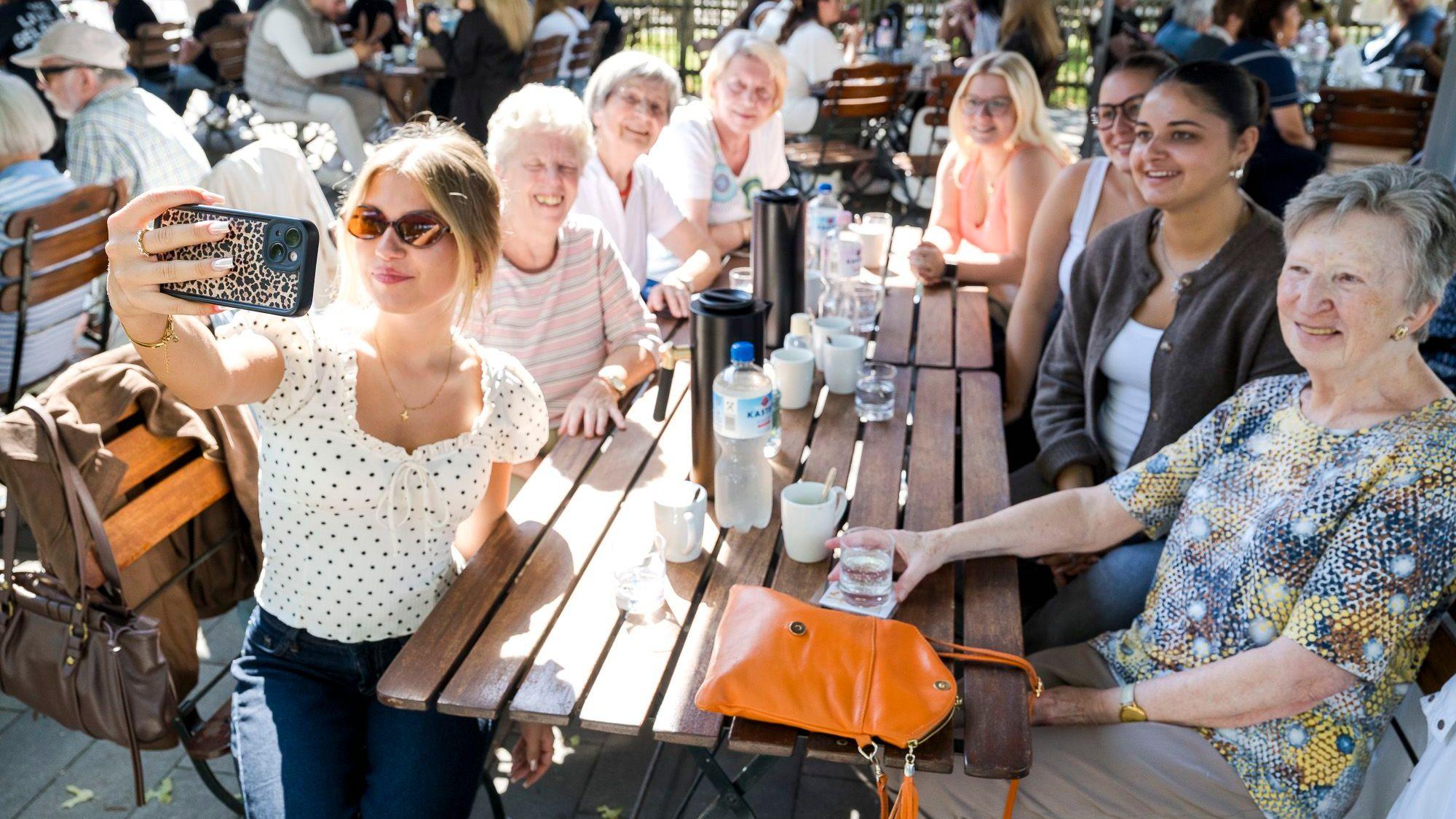 Junge und alte Frauen sitzen um einen Tisch im Gartenrestaurant; eine junge Frau nimmt ein Selfie mit der Gruppe auf.