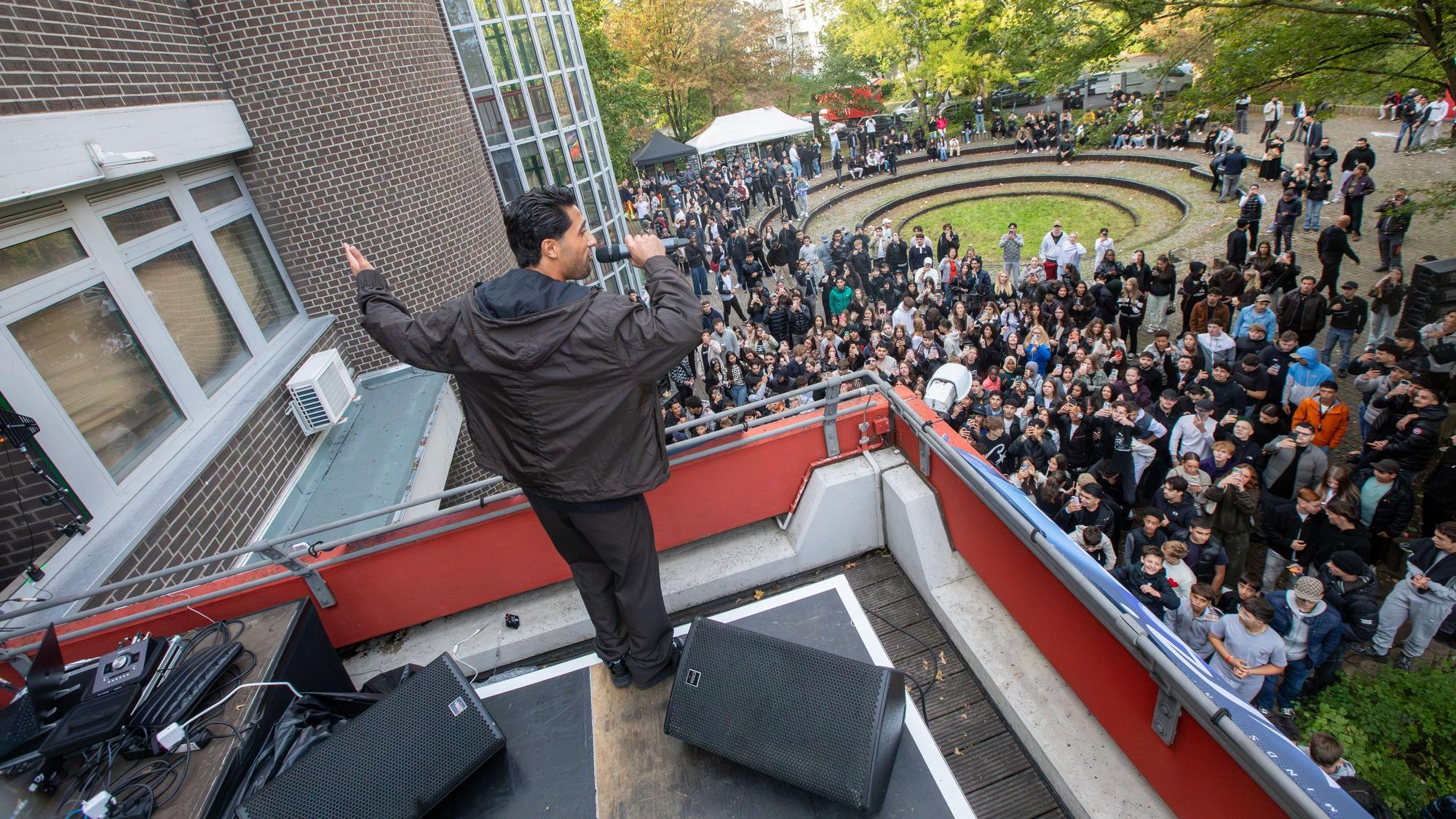 Ein junger Mann singt auf einer Dachterrasse. Unten auf dem Rasen steht eine jubelnde Menge.