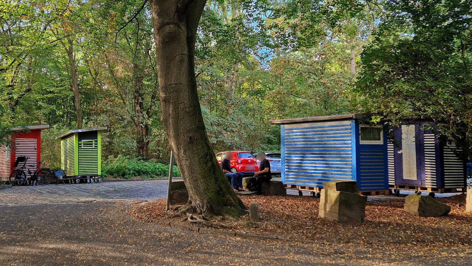 Auf einem Parkplatz in einem Waldgebiet stehen vier bunte Holzhütten.