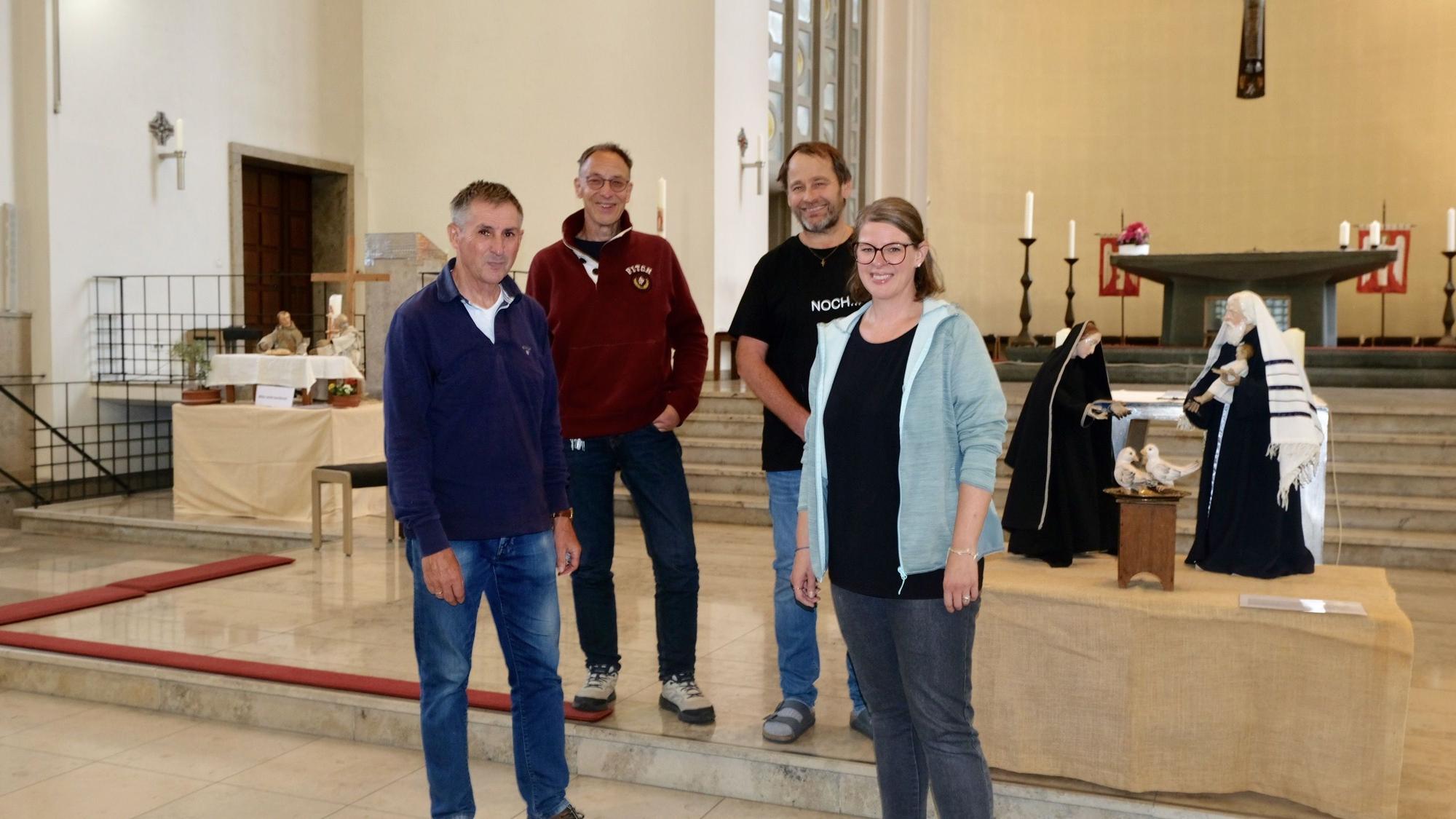Drei Männer und eine Frau stehen im Altarraum einer Kirche, wo verschiedene Krippenfiguren aufgebaut sind.