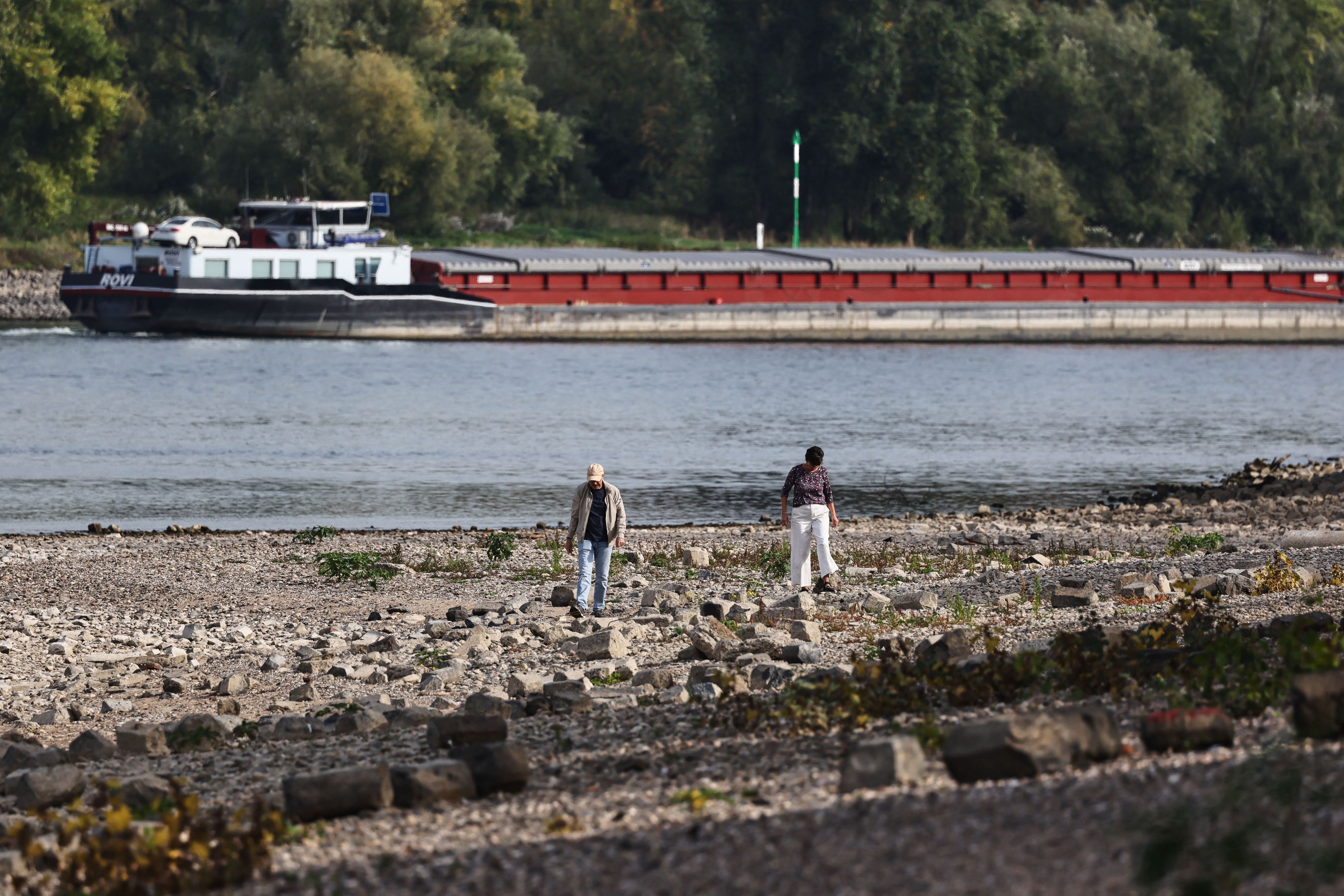 Spaziergänger gehen am Rheinufer in Köln entlang. Im Hintergrund fährt ein Binnenschiff.