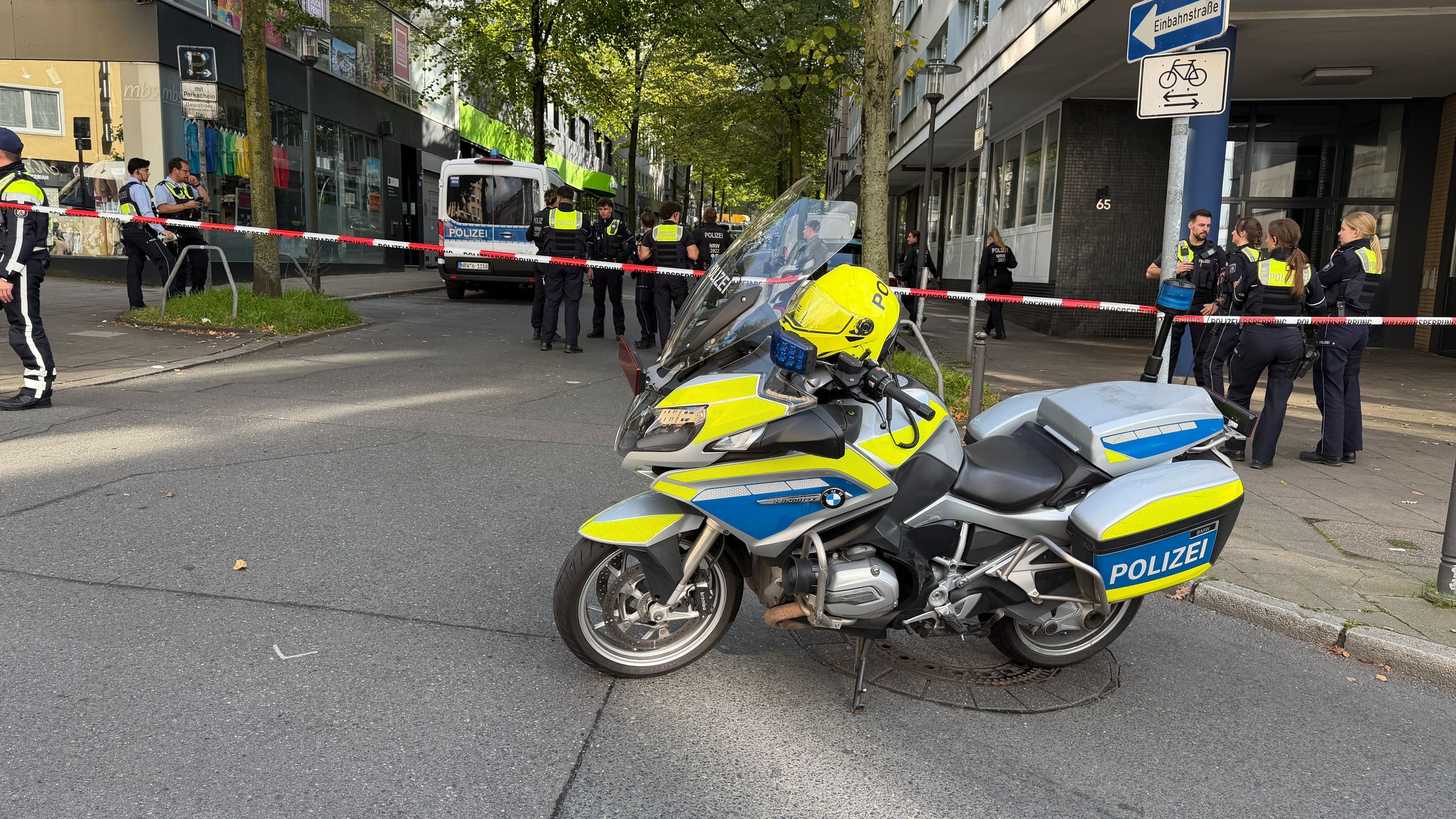 Absperrband hinter einem Polizeimotorrad in der Nähe eines Tatorts. Einsatzkräfte stehen auf einem Parkplatz. Zwei Unbekannte haben mitten in Essen zwei Justizbeamte angegriffen und einen 34 Jahre alten Häftling befreit.