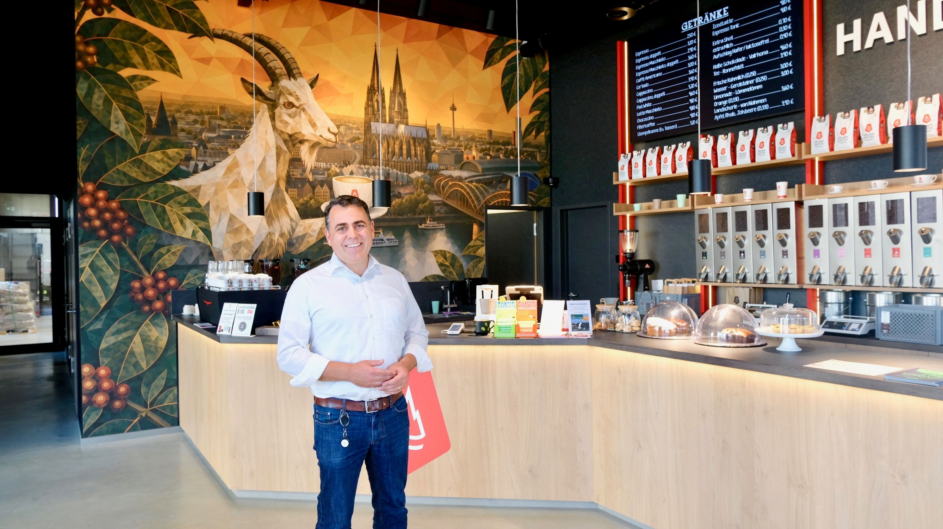 Ein Mann im weißen Hemd steht vor einem großen Verkaufstresen, hinter ihm ziert eine große Fototapete mit Dom und Geißbock die Wand