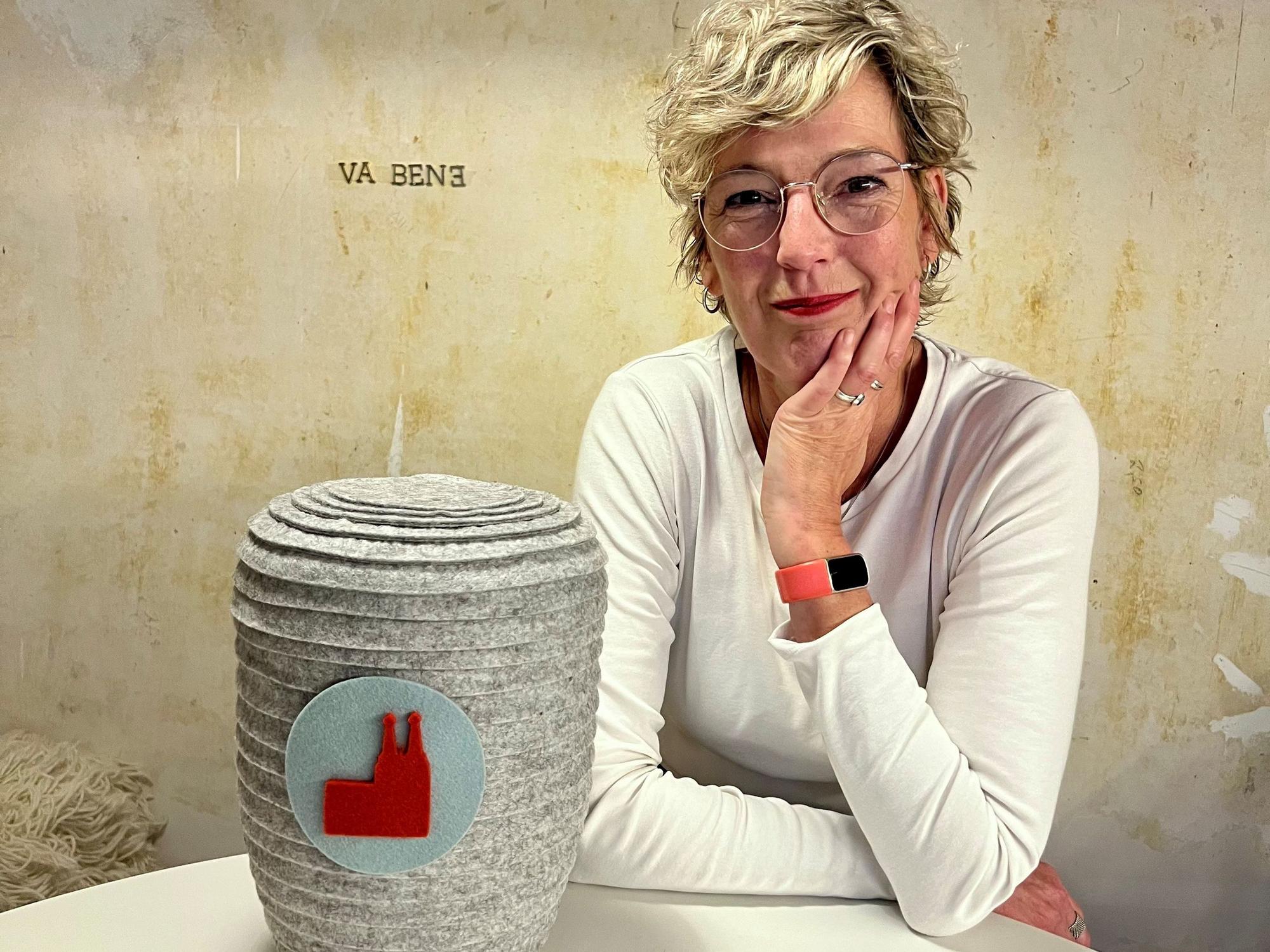 Eine Frau im weißen T-Shirt sitzt an einem Tisch, vor ihr steht eine Urne im Filzmantel.