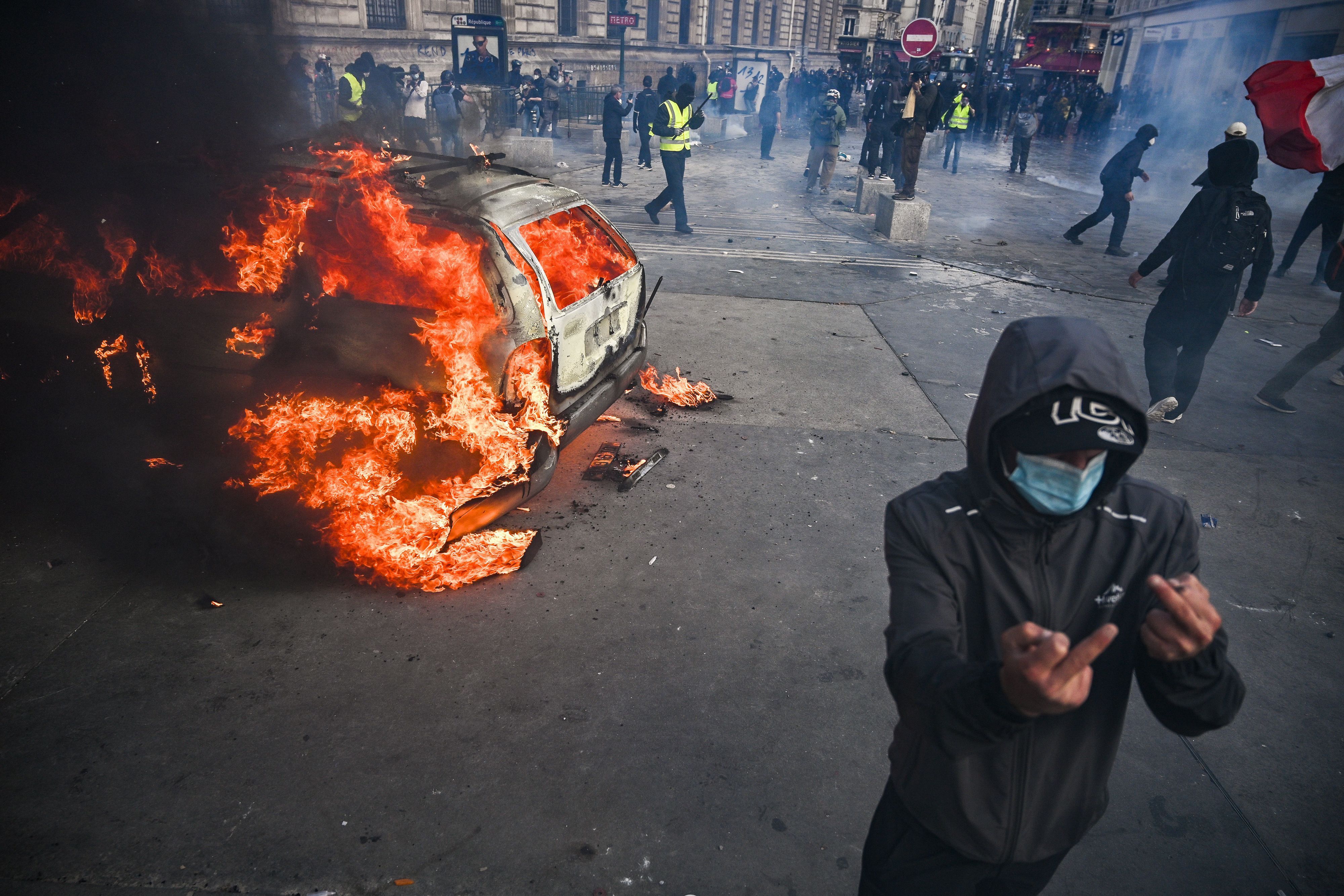 Auf dem Place de la République brennt ein Auto, davor zeigt eine vermummte Person den Mittelfinger.