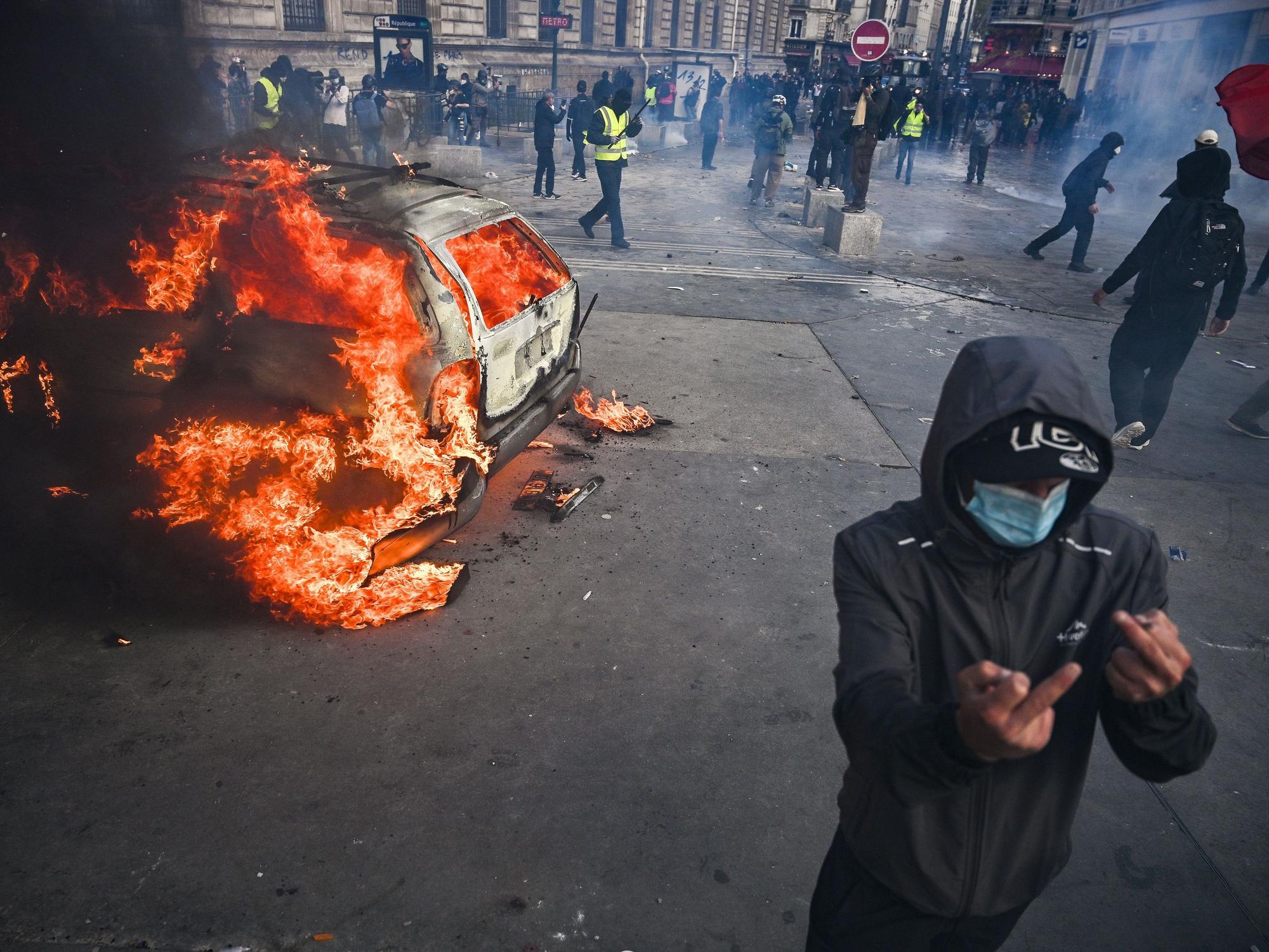 Auf dem Place de la République brennt ein Auto, davor zeigt eine vermummte Person den Mittelfinger.