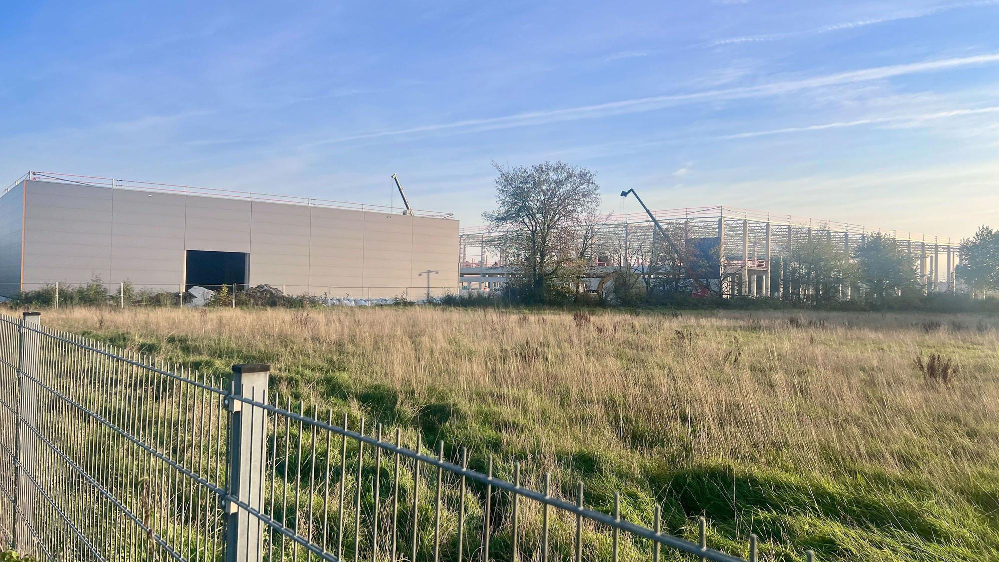 Hinter einem Metallzaun und einer hohen Wiese stehen links eine fertige Industriehalle mit heller Fassade und rechts eine im Bau befindliche Halle aus Stahlgerüst mit Baukränen unter blauem Himmel.