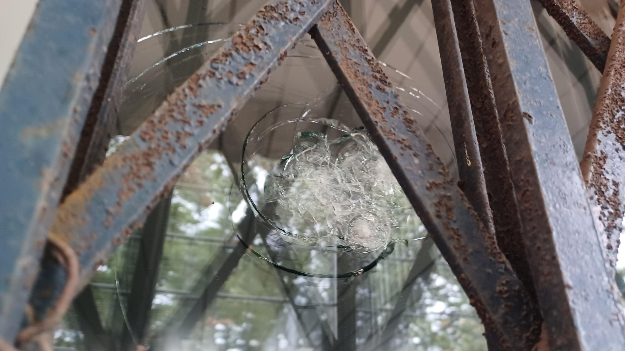 Zu sehen ist eine Glasscheibe mit einem großen Einschlagloch und radialen Sprüngen, eingefasst von verrosteten Metallstreben.