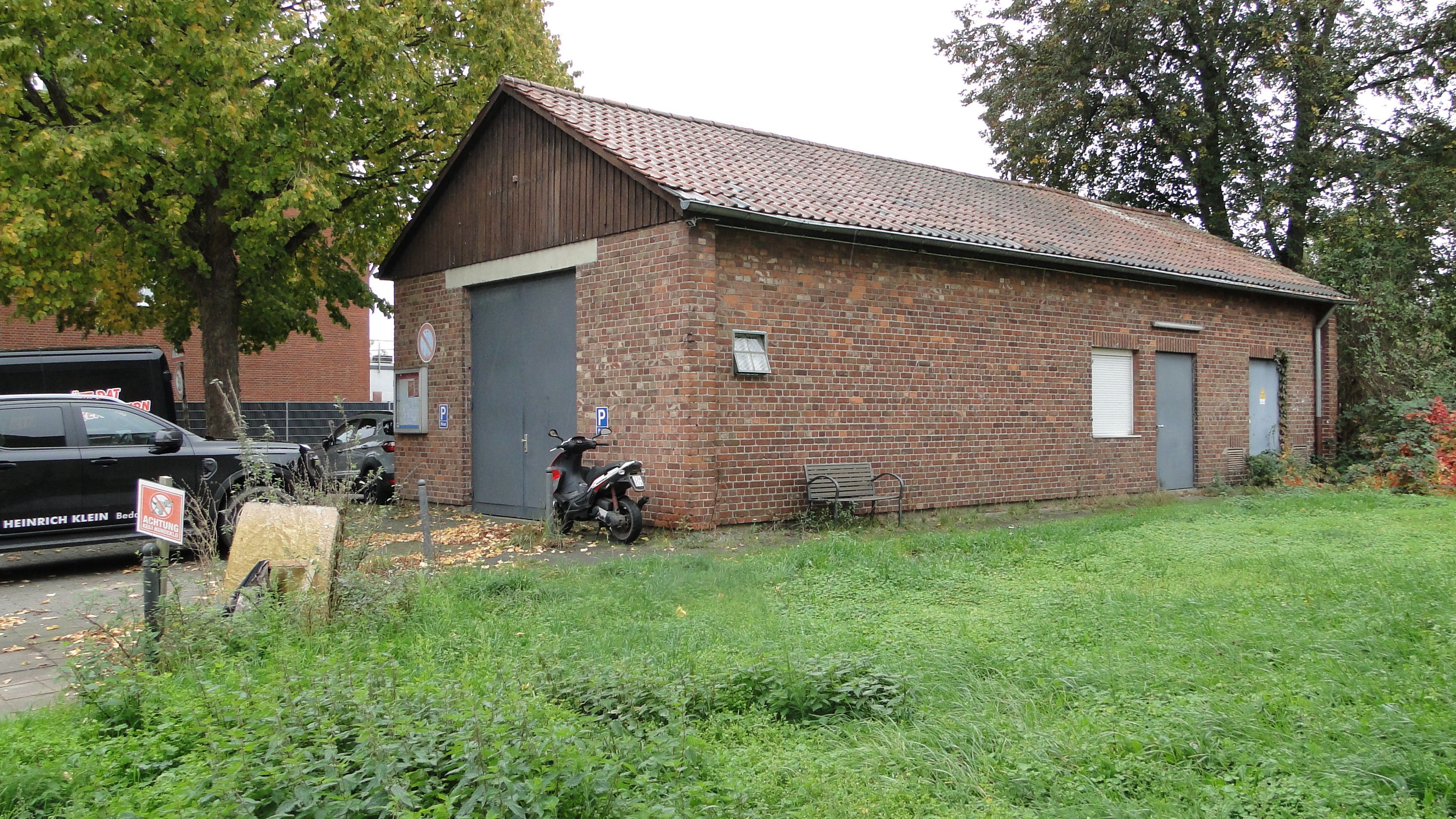 Ein niedriges rotes Backsteingebäude mit Ziegeldach und grauem Rolltor steht an einer Straße. Davor parken ein Motorroller und ein Pkw und vor dem Gebäude befindet sich eine Wiese.