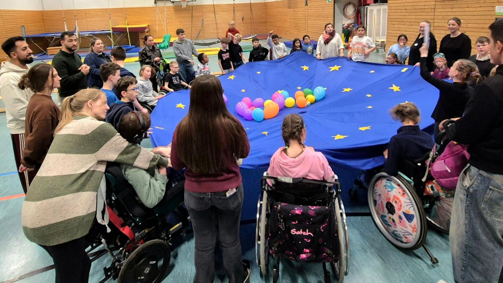 Um ein blaues Tuch herum haben sich Kinder in Rollstühlen versammelt. Sie halten es gemeinsam hoch, in der Mitte des Tuchs liegen bunte Luftballons.