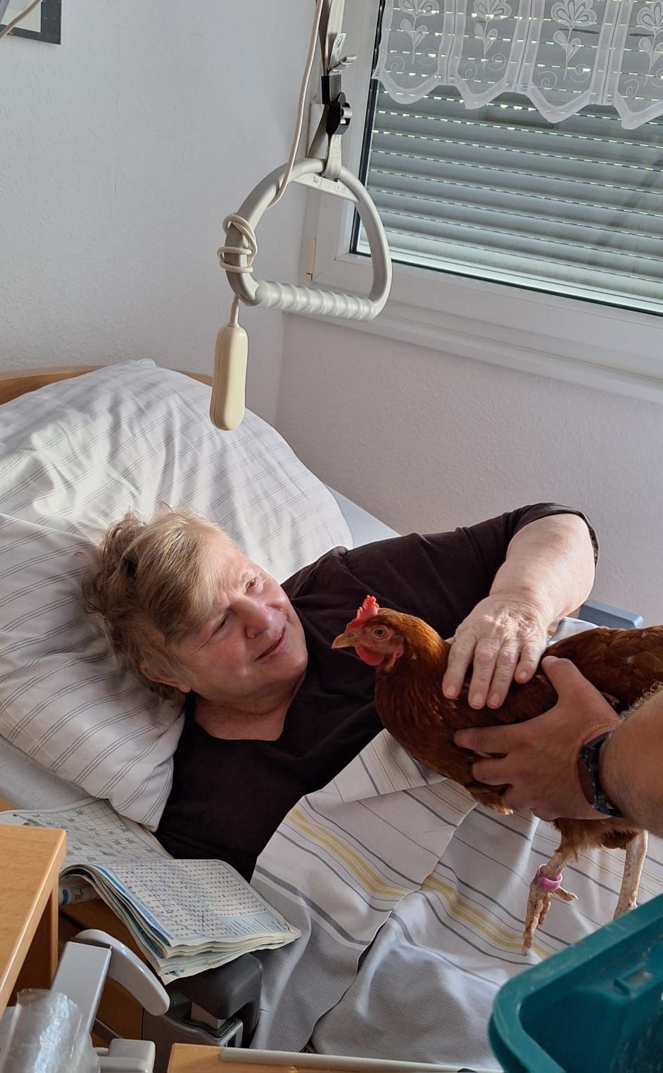 Eine alte Frau liegt im Bett und streichelt vorsichtig ein Huhn, das ihr hingehalten wird.
