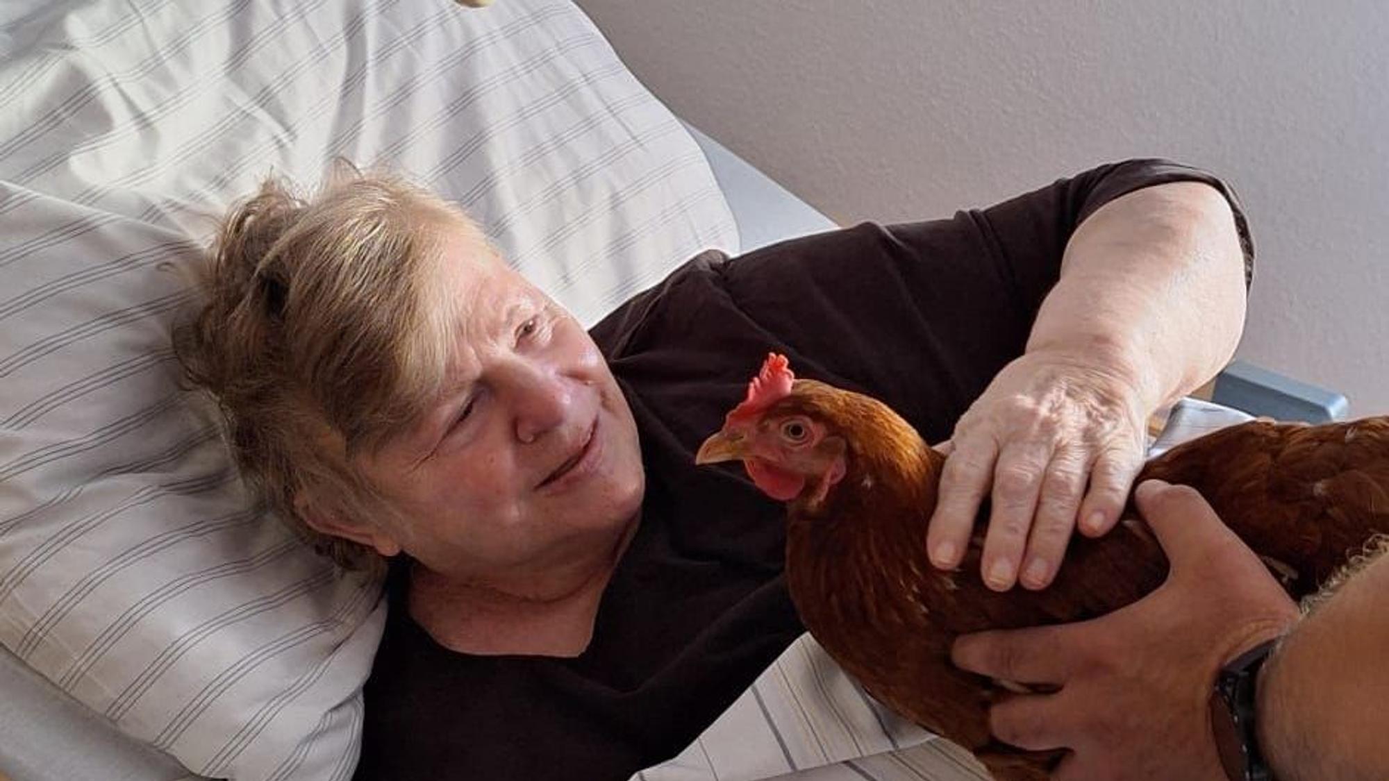Eine alte Frau liegt im Bett und streichelt vorsichtig ein Huhn, das ihr hingehalten wird.