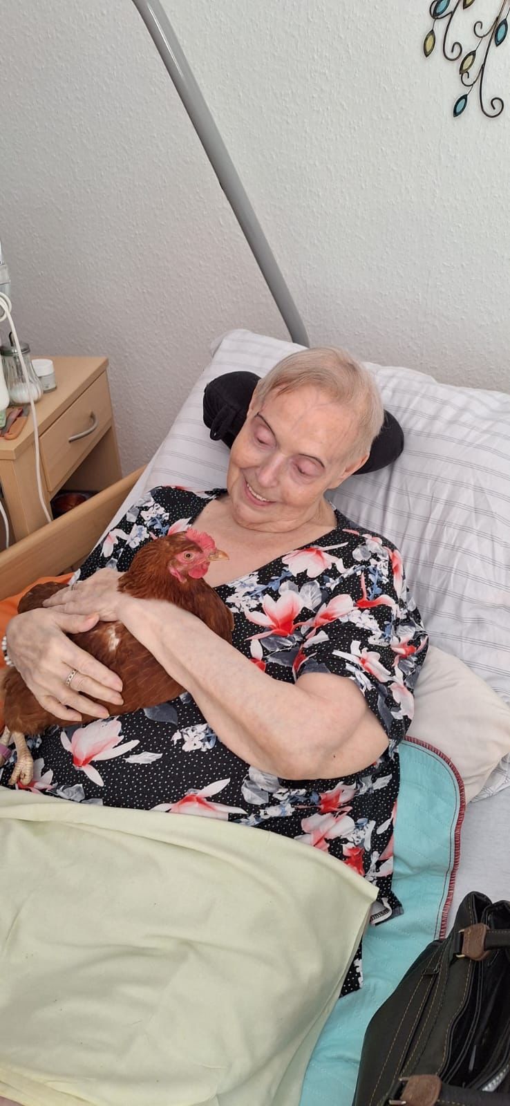 Eine alte Frau liegt im Bett und lächelt glücklich ein Huhn an, das sie im Arm hält.