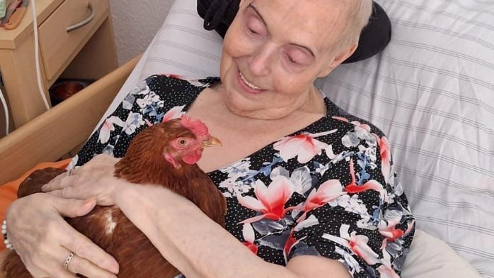 Eine alte Frau liegt im Bett und lächelt glücklich ein Huhn an, das sie im Arm hält.