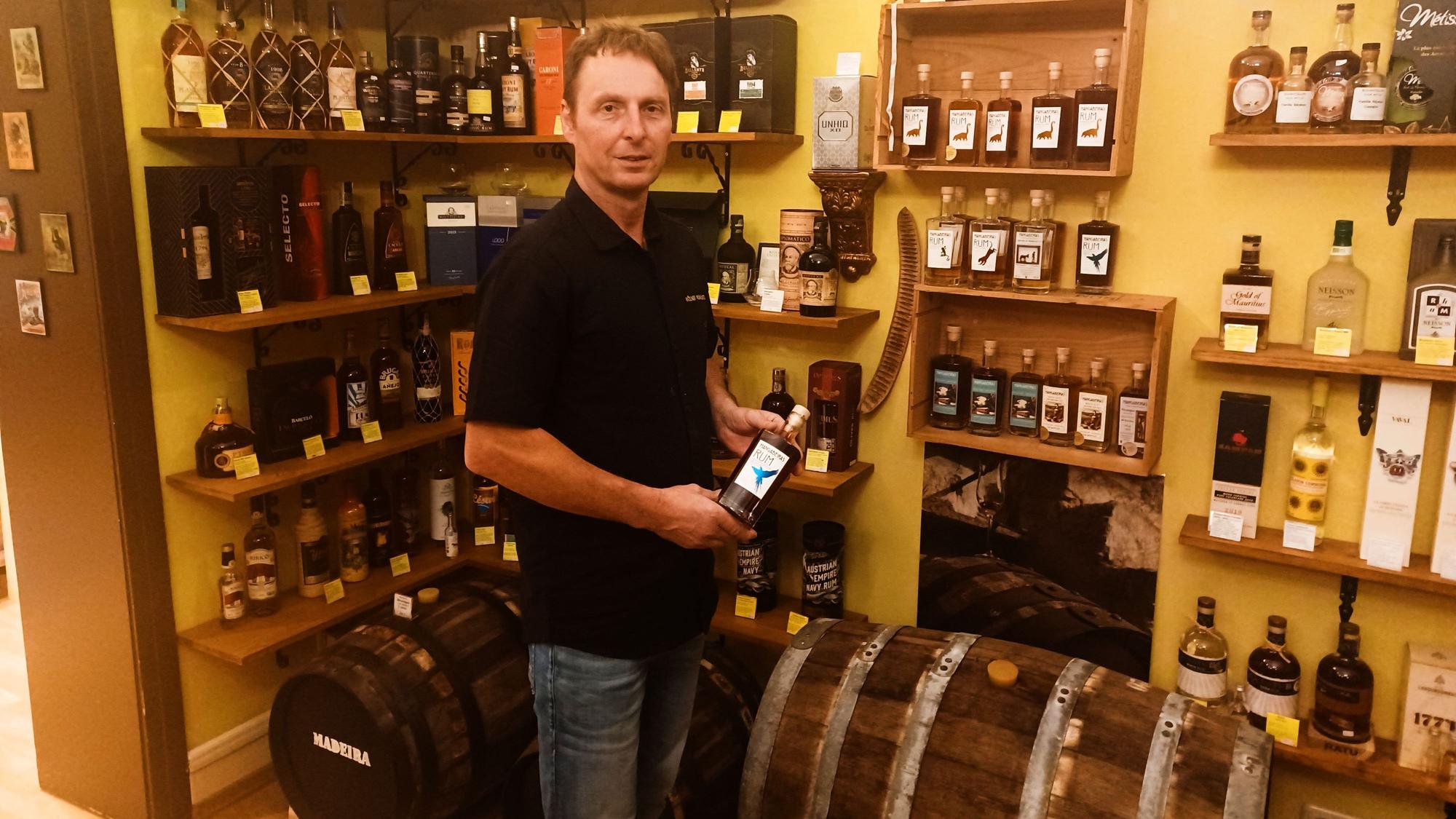 Ein Mann im schwarzen Shirt und Jeans steht in einem Laden voller Spirituosen-Flaschen. In der Hand hält er eine Flasche.