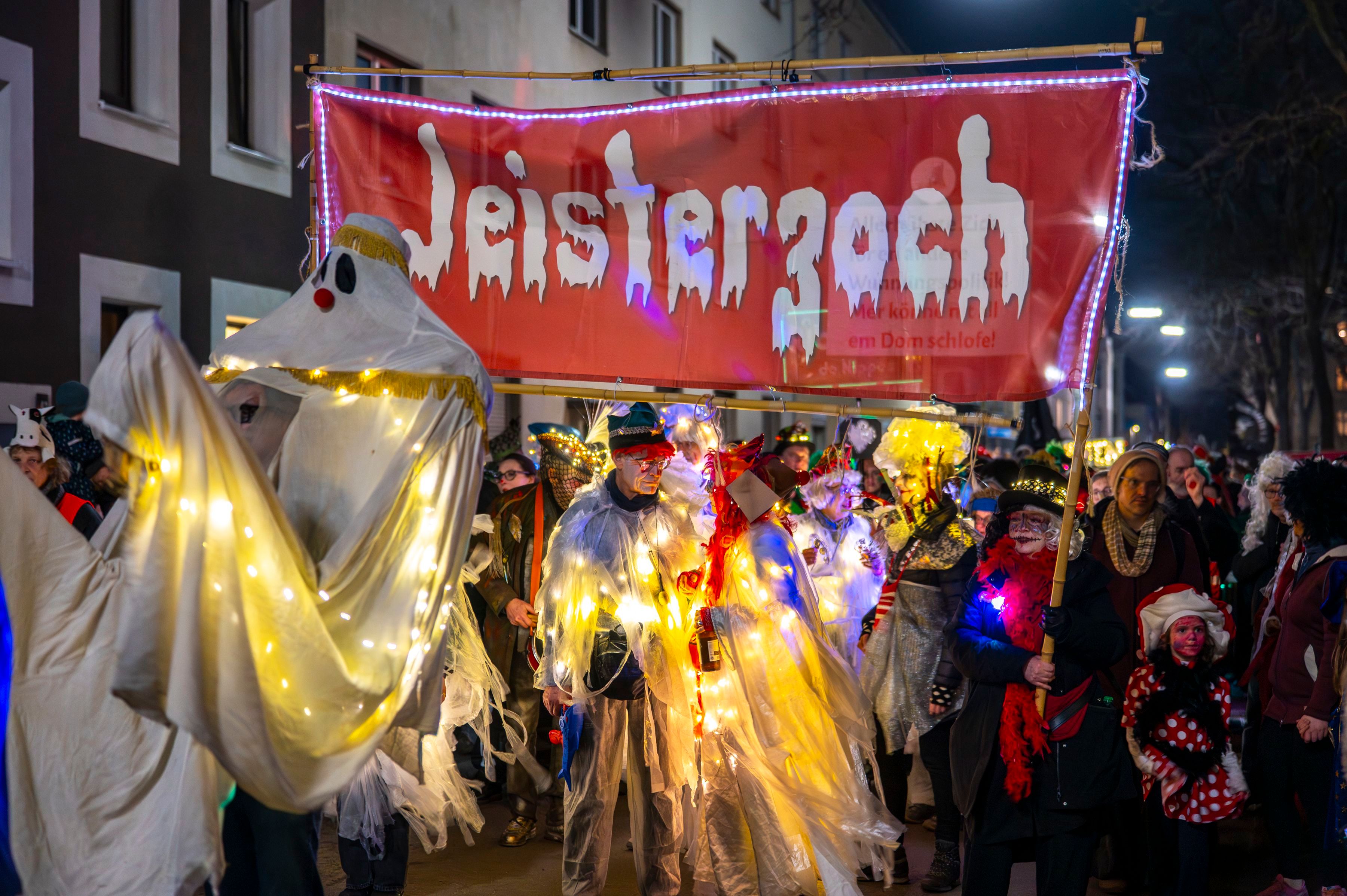 Verkleidete Gespenster stehen auf der Straße. Sie halten ein Banner mit der Aufschrift „Jeisterzoch “(Geisterzug) hoch.