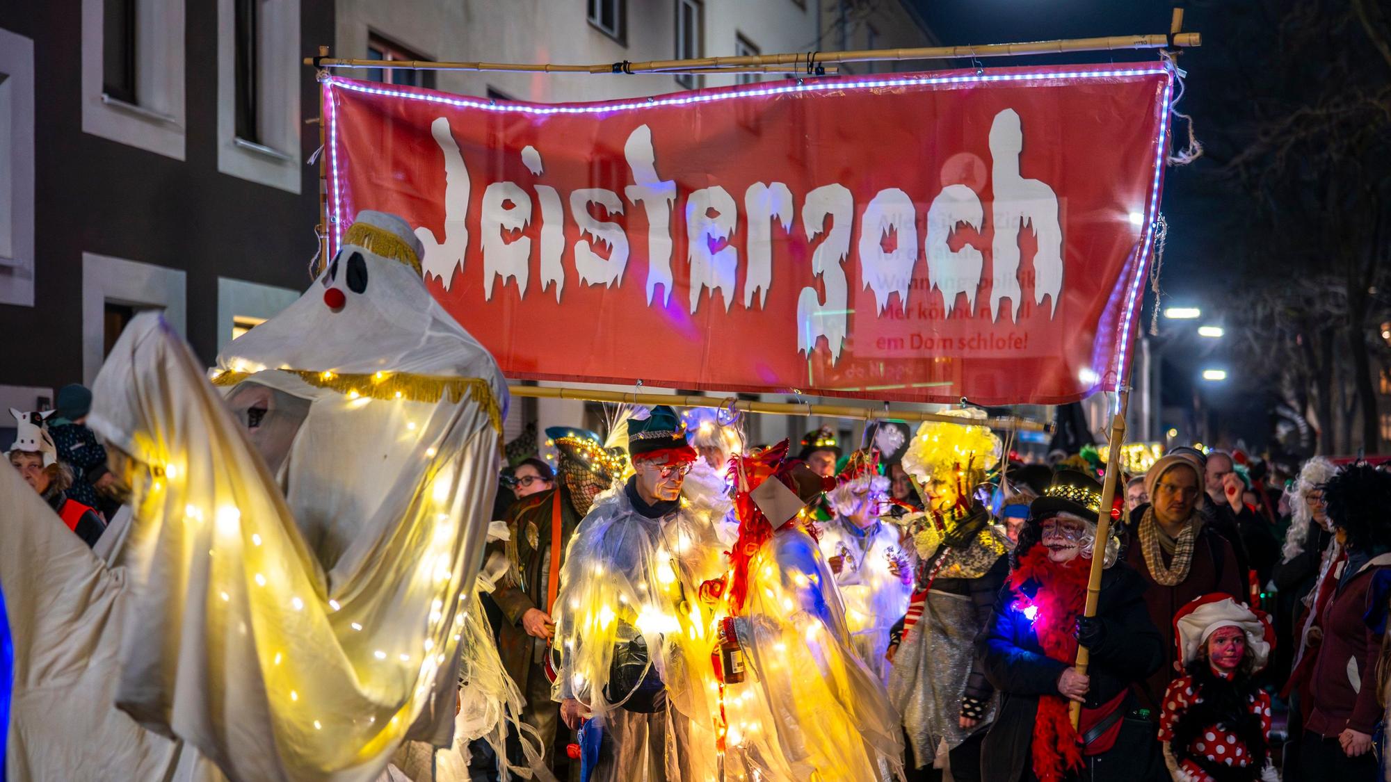 Verkleidete Gespenster stehen auf der Straße. Sie halten ein Banner mit der Aufschrift „Jeisterzoch “(Geisterzug) hoch.