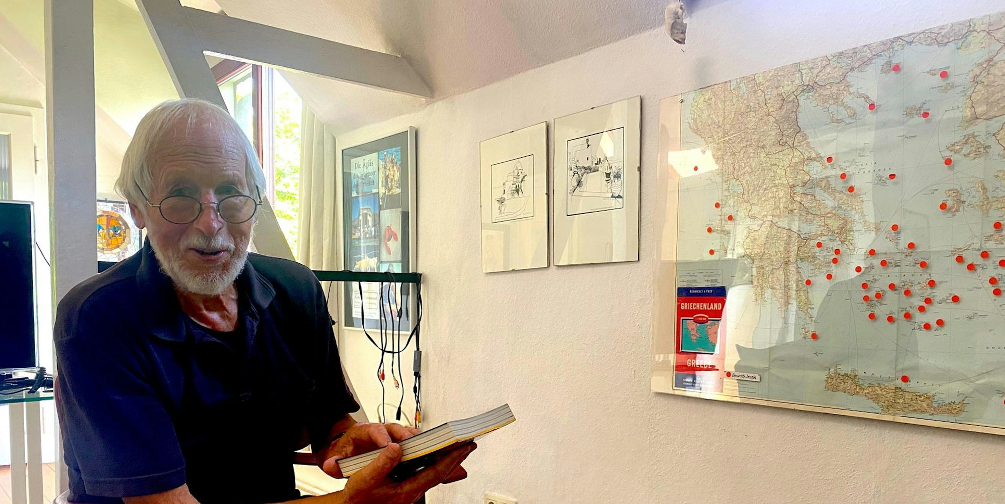 Kurt Schreiner vor einer Weltkarte mit vielen Pins an den bereisten Orten. In der Hand hält er eines von seinen selbstgeschriebenen Reisebüchern.