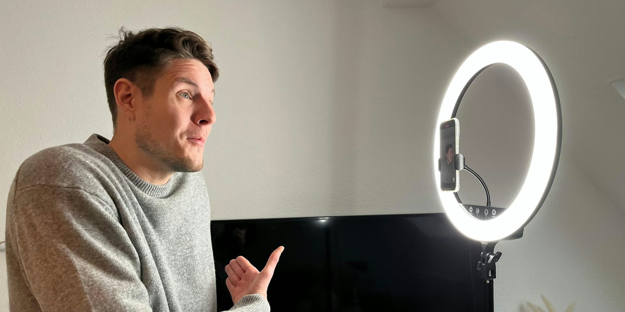 Ein Mann im grauen Pulli, mit braunen Haaren und Dreitagebart steht vor einem Ringlicht und spricht in die Handykamera.