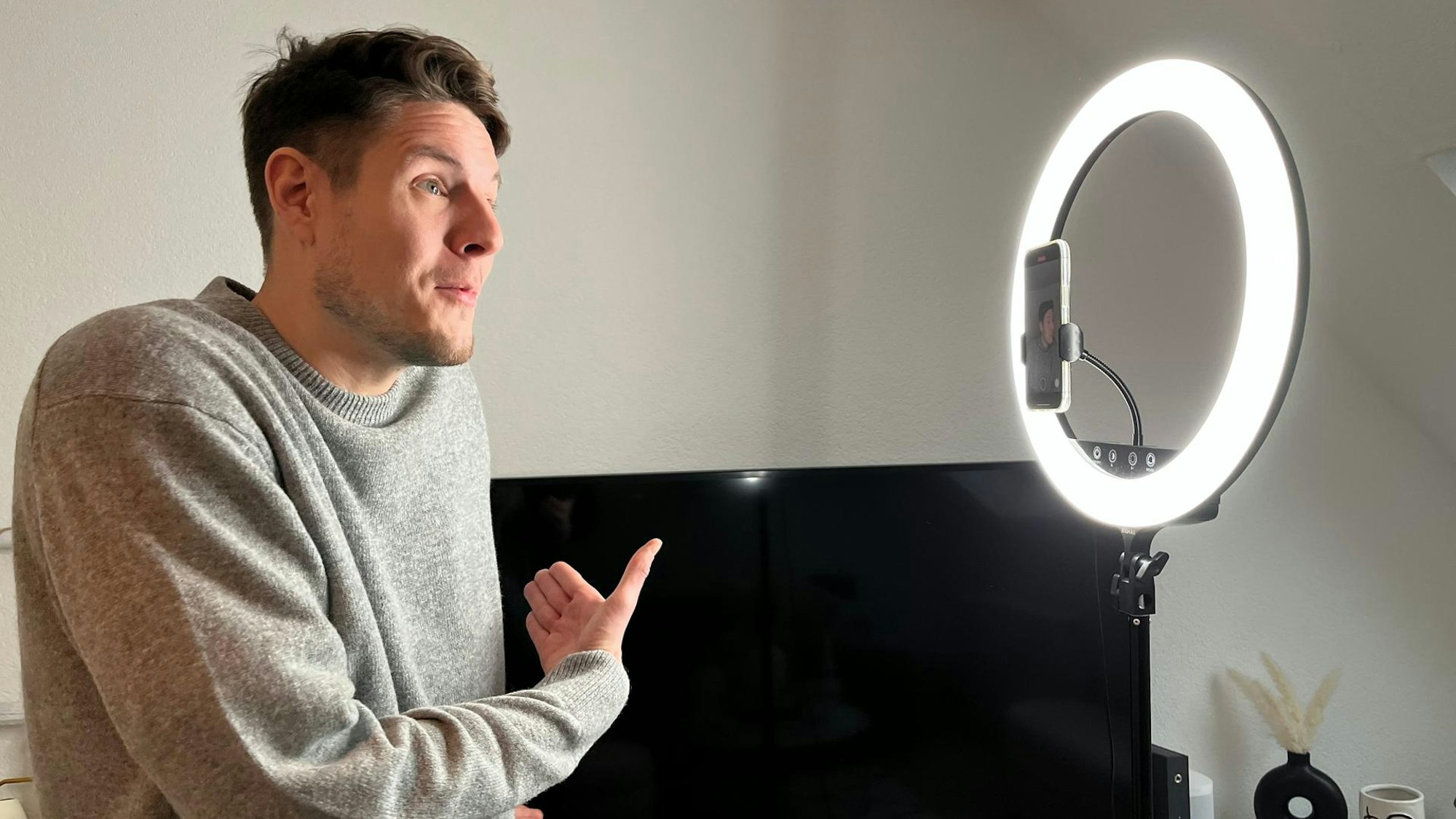Ein Mann im grauen Pulli, mit braunen Haaren und Dreitagebart steht vor einem Ringlicht und spricht in die Handykamera.