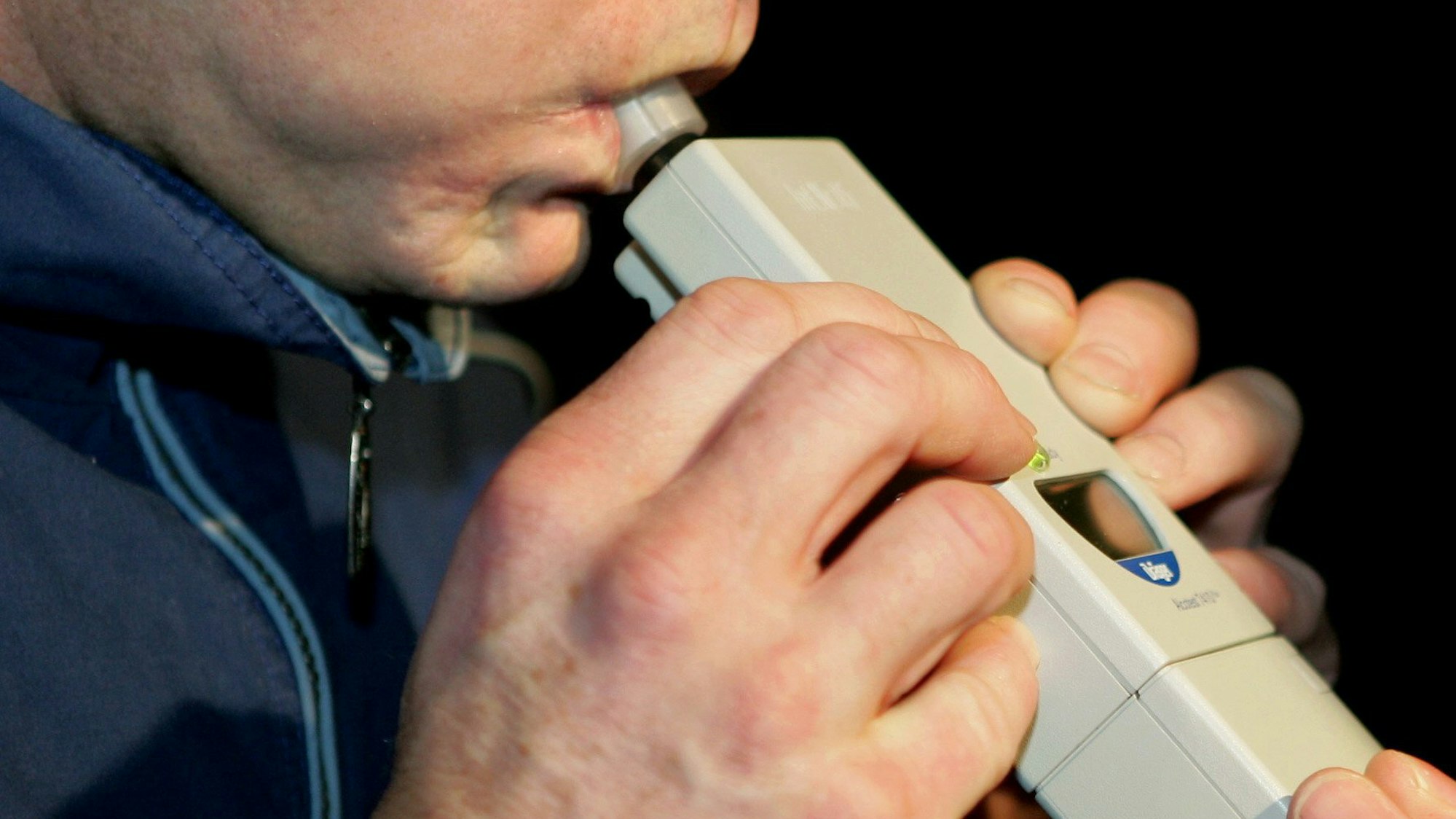 Das Symbolfoto zeigt die Hände und die untere Gesichtshälfte eines Mannes, der in ein Alkoholtestgerät pustet.