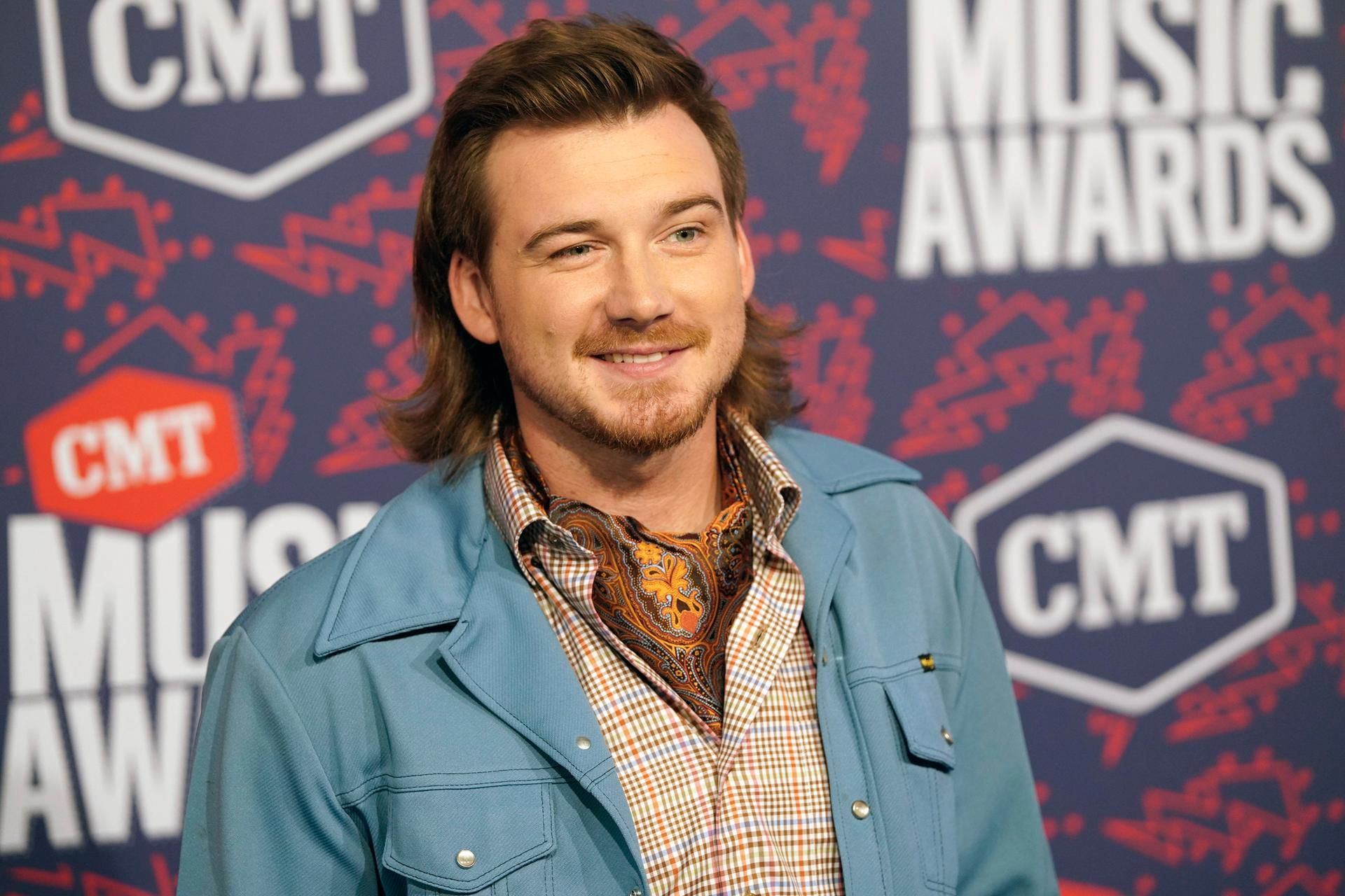 Morgan Wallen mit Vokuhila, babyblauer Stoffjacke und Halstuch. Er grinst smart in die Kamera.