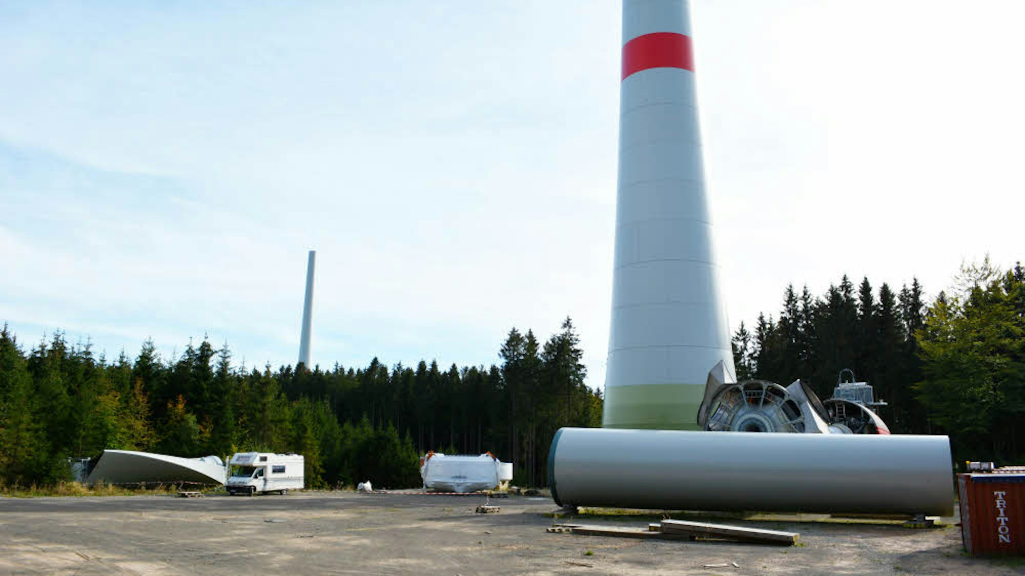 Der untere Teil einer Windkraftanlage mit einem roten Streifen ist zu sehen. Am Boden liegen weitere Bauteile.