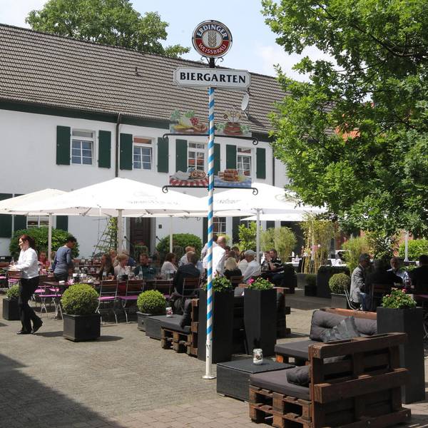 Vor einem weißen Haus stehen Tische und Stühle unter Sonnenschirmen. Ein Biergarten-Schild steht auf dem Platz.