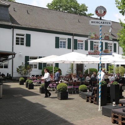 Vor einem weißen Haus stehen Tische und Stühle unter Sonnenschirmen. Ein Biergarten-Schild steht auf dem Platz.