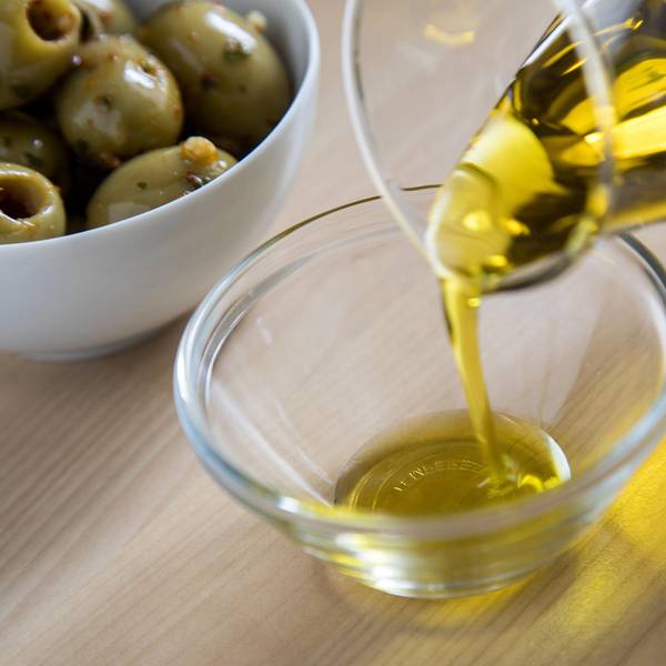 Aus einer Glaskaraffe wird Olivenöl in eine Glasschale gegossen, im Hintergrund eine Porzellanschale mit grünen Oliven.