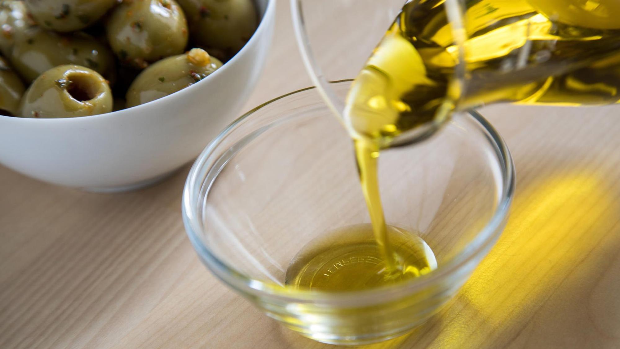 Aus einer Glaskaraffe wird Olivenöl in eine Glasschale gegossen, im Hintergrund eine Porzellanschale mit grünen Oliven.
