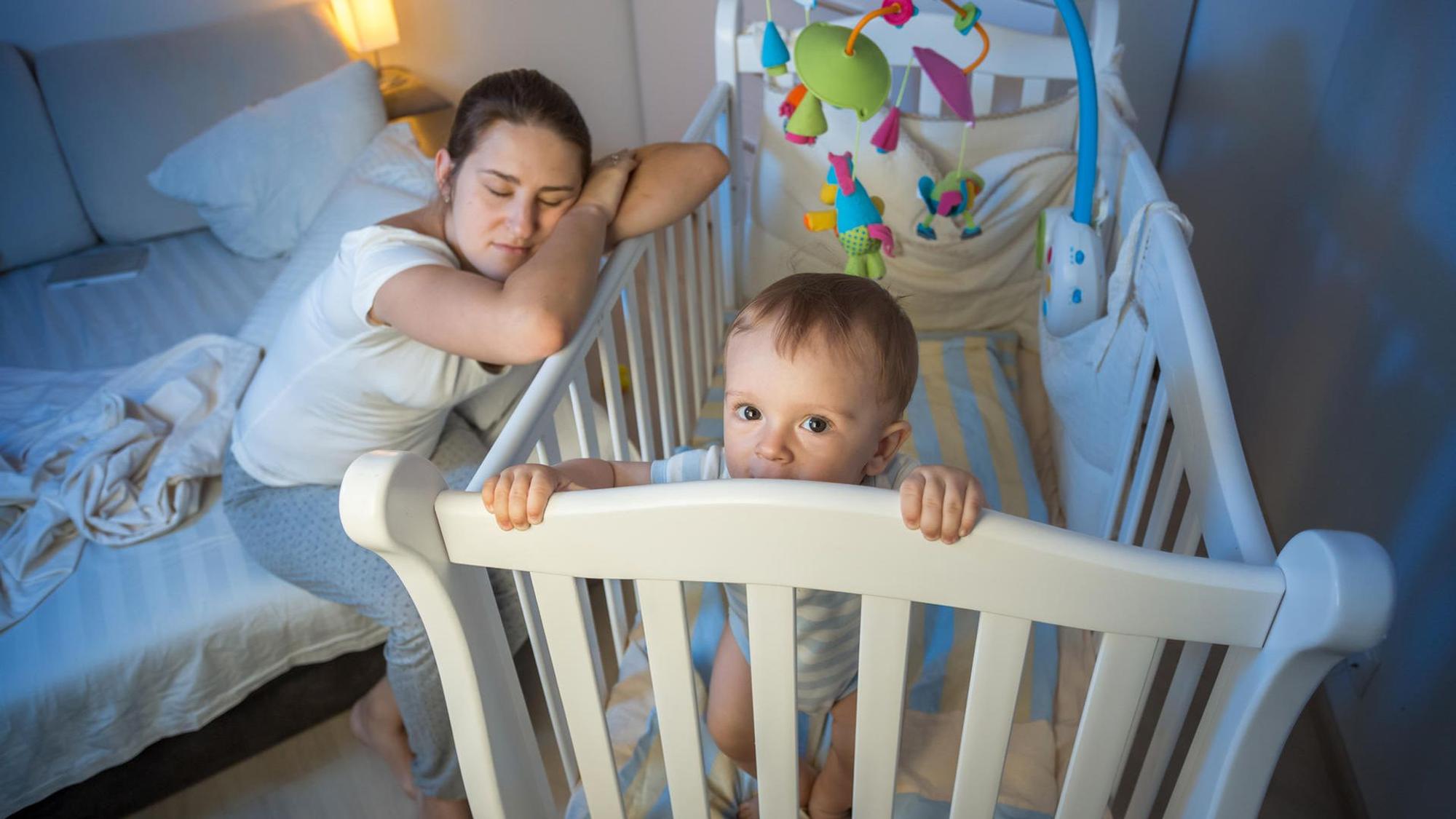 Eine Mutter schläft, an die Wiege des wachen Babys gelehnt.