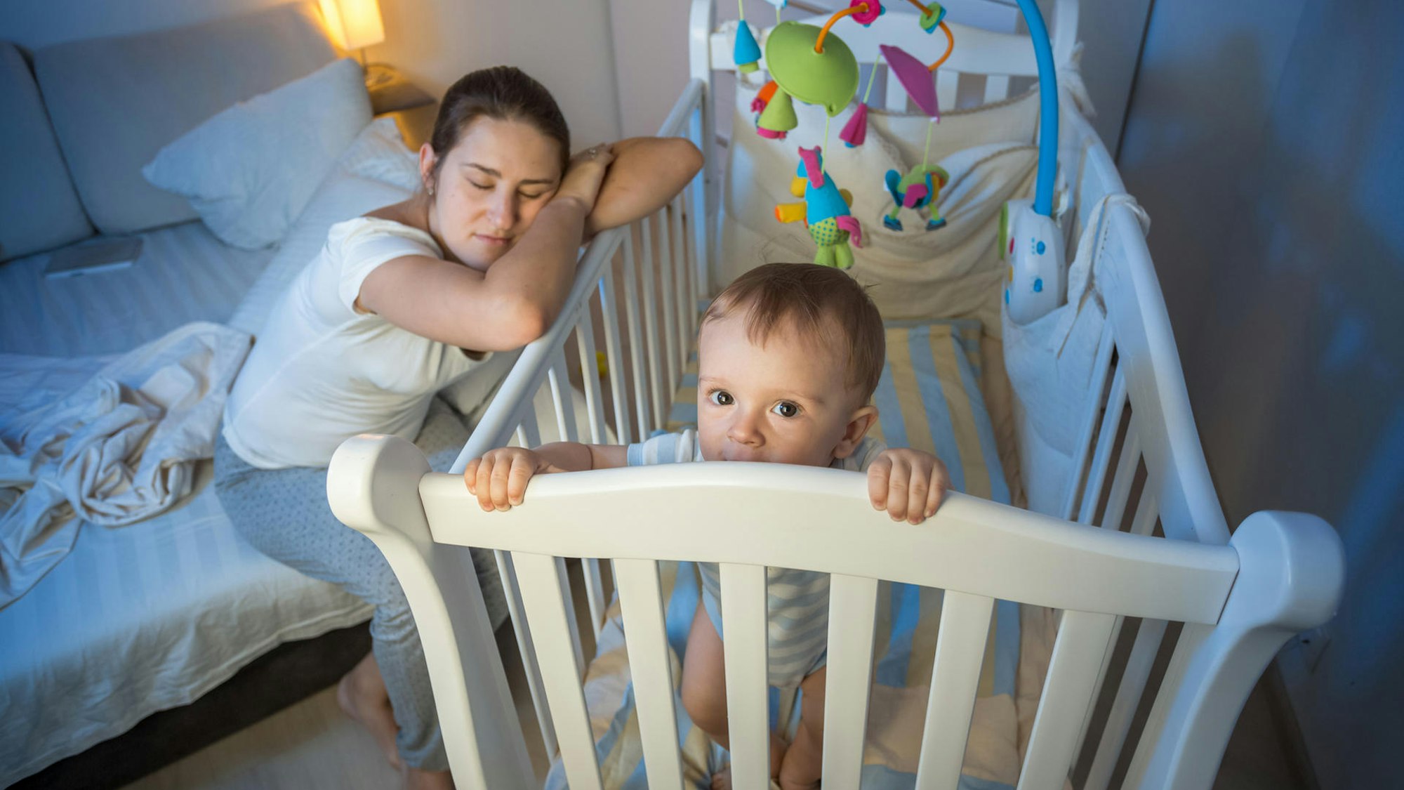 Eine Mutter schläft, an die Wiege des wachen Babys gelehnt.