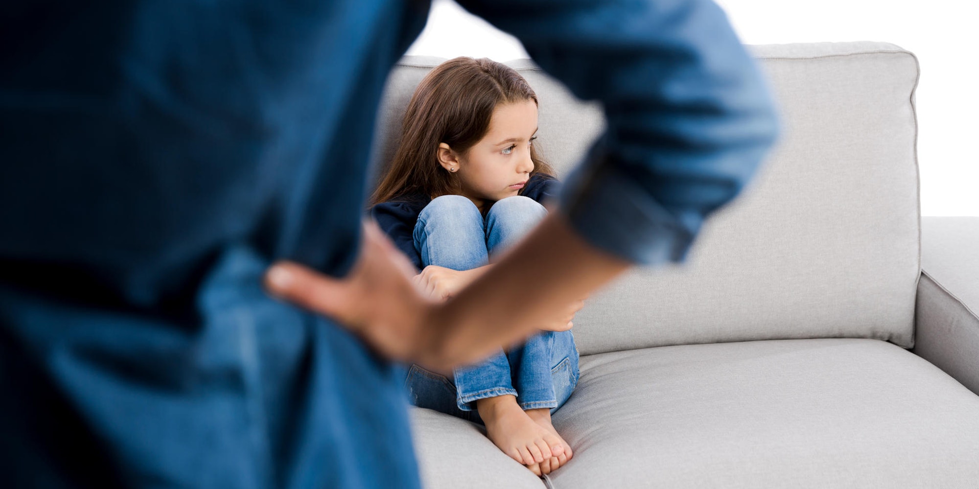 Mädchen sitzt auf der Couch und dreht sich weg, ein Erwachsenen von hinten, der die Hände in die Hüften stützt.