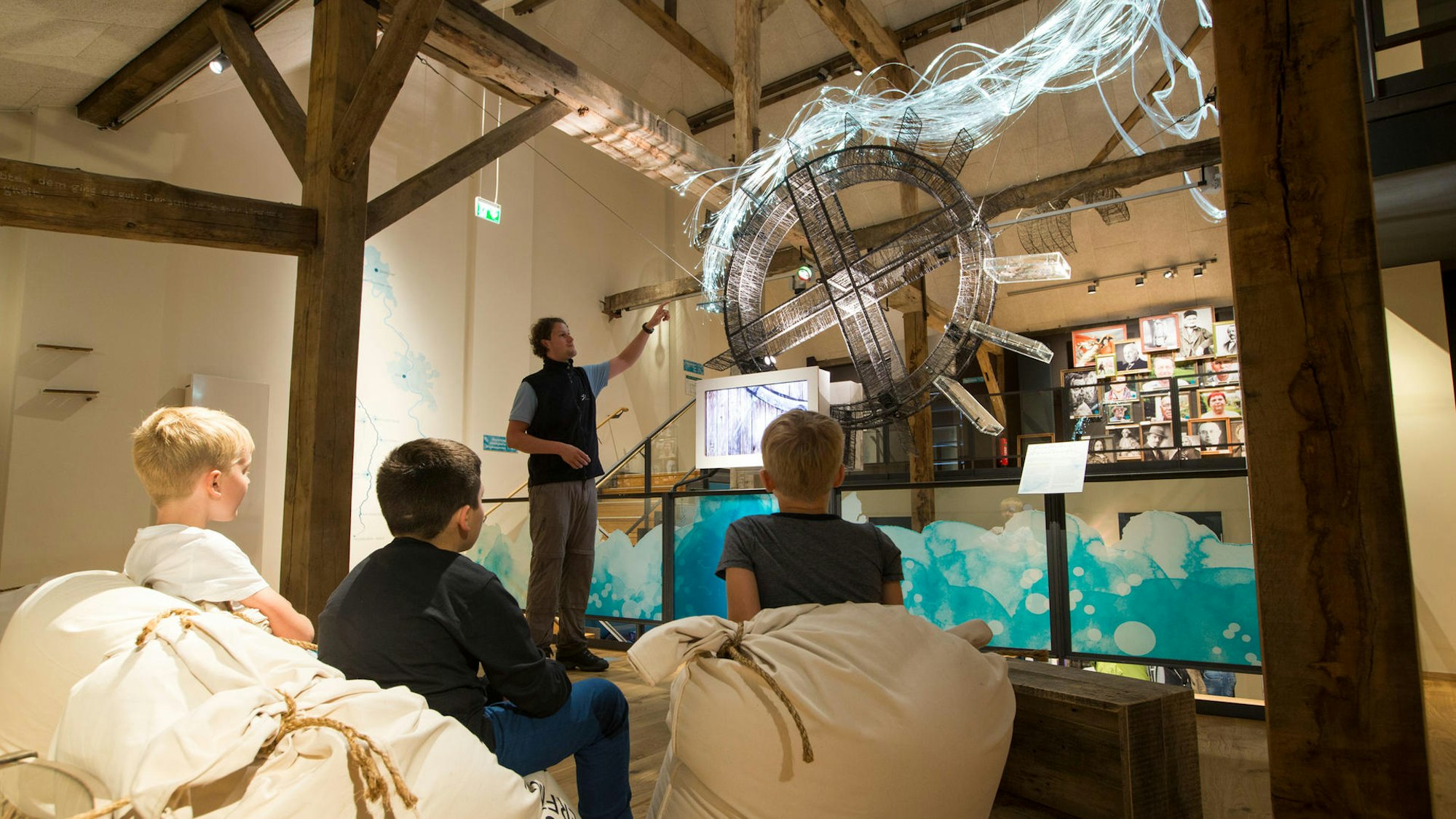 drei Kinder sitzen vor einem Wasserrad, an dem ein Mann etwas zeigt