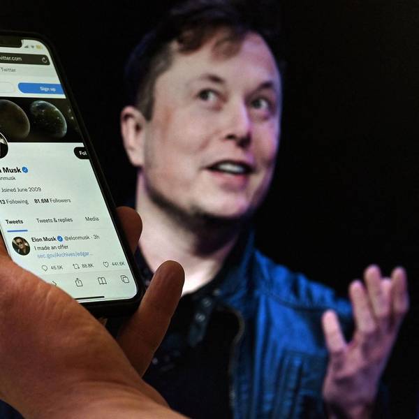 Elon Musk mit erhobenen Händen beim Reden hinter einem Handy, dass sein Twitter-Profil zeigt.