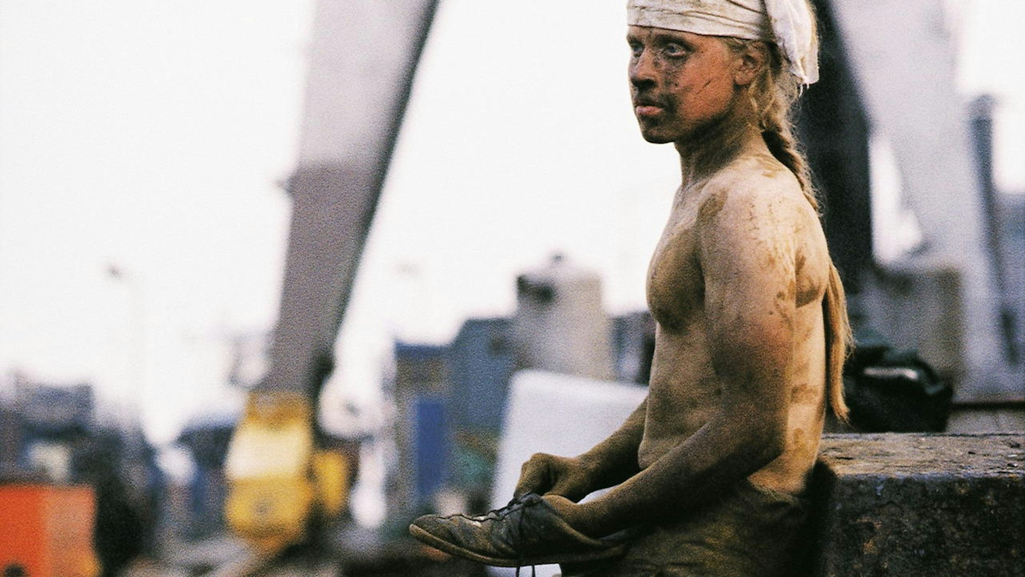 Ein junger Mann steht mit Schmutz überall gegen einen Betonklotz gelehnt, und hat einen Schuh in der Hand.