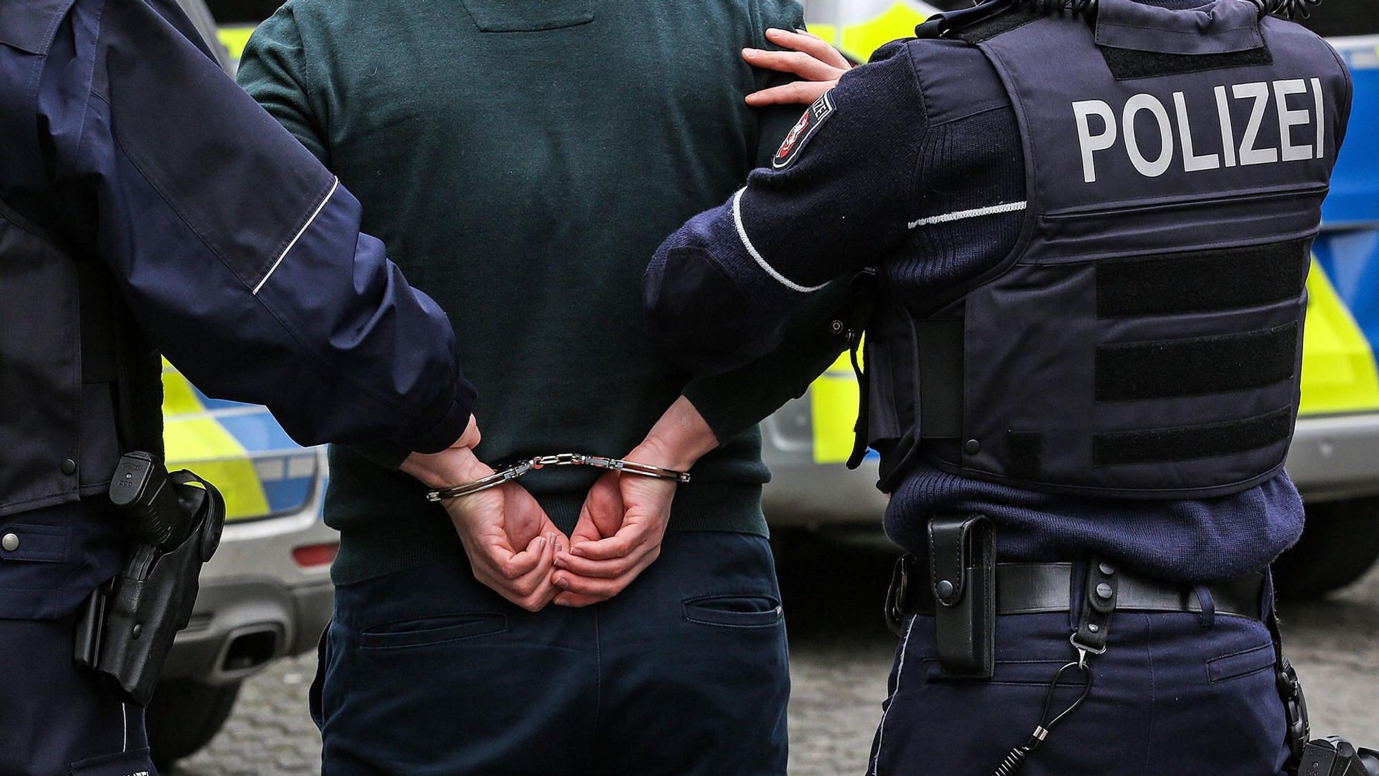 Das Symbolbild (Detailaufnahme) zeigt zwei Polizisten von hinten, die einen Mann in Handschellen abführen.
