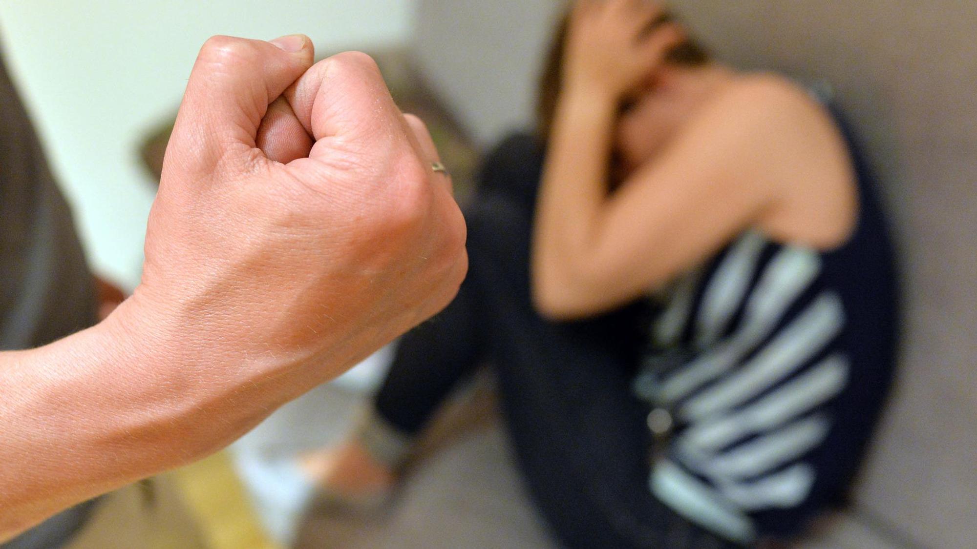 Im Vordergrund eine geballte Faust, unscharf im Hintergrund eine zusammengekauerte Frau am Boden.