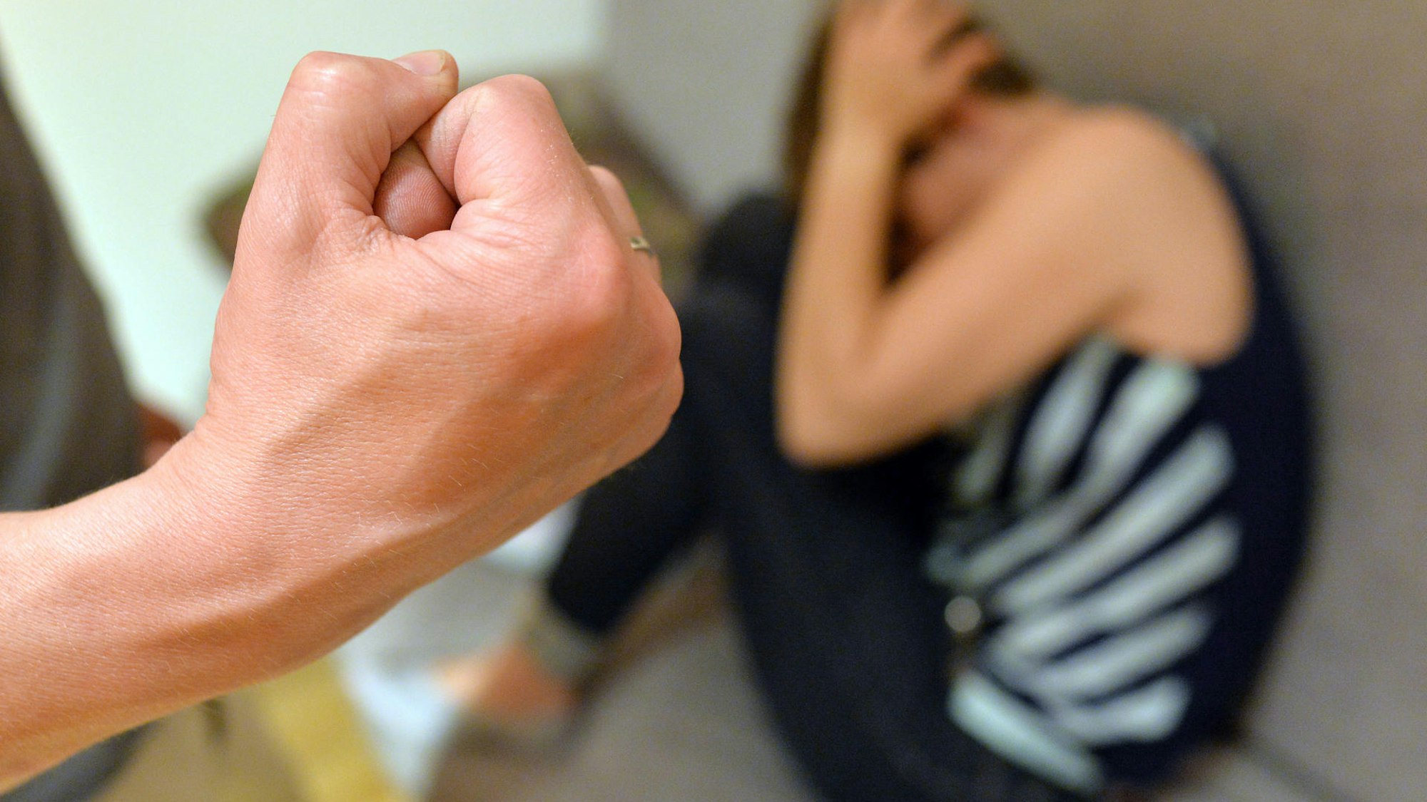 Im Vordergrund eine geballte Faust, unscharf im Hintergrund eine zusammengekauerte Frau am Boden.